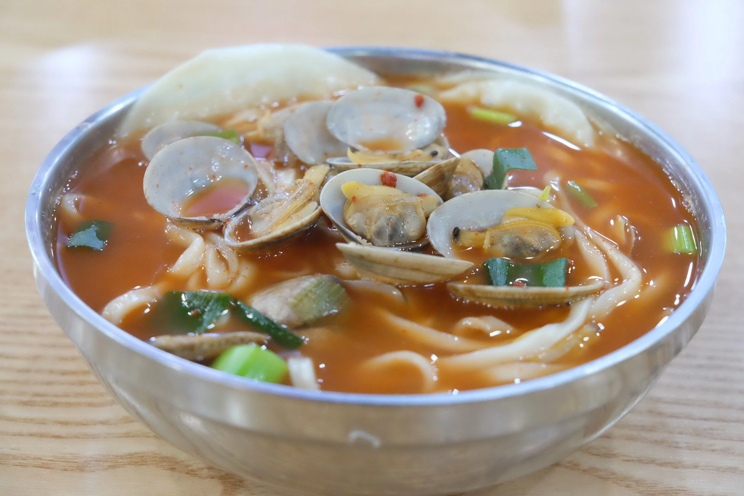 A bowl of shacha seafood noodle soup in Fujian, China with clams, green onions, and chili in a red broth on a wooden table.