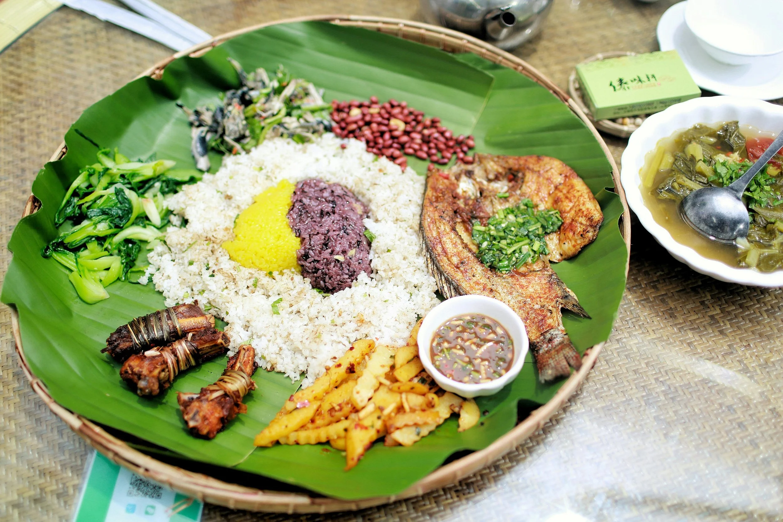 A traditional Yunnan ethnic meal served on a large banana leaf with rice, vegetables, fish, grilled meat, and dipping sauces.