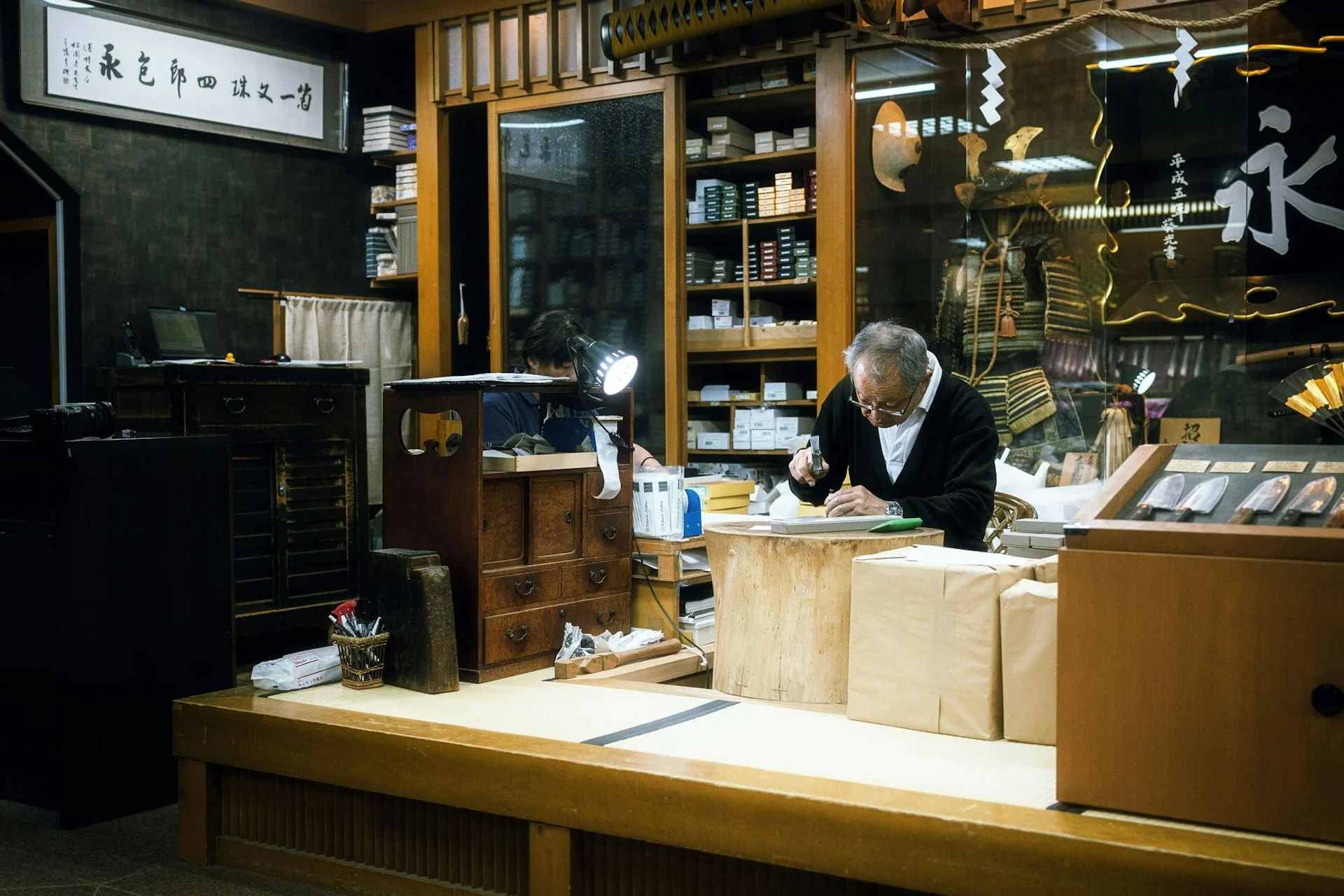 Knife maker working at the counter of a traditional Japanese knife shop, with swords and blades on display behind