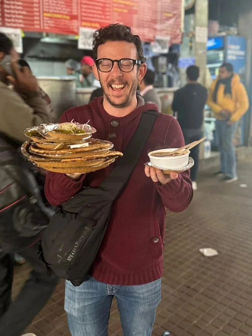 A man with glasses in a maroon sweater is smiling and holding a stack of plates with food in one hand and a bowl with a spoon in the other at a busy outdoor food market.