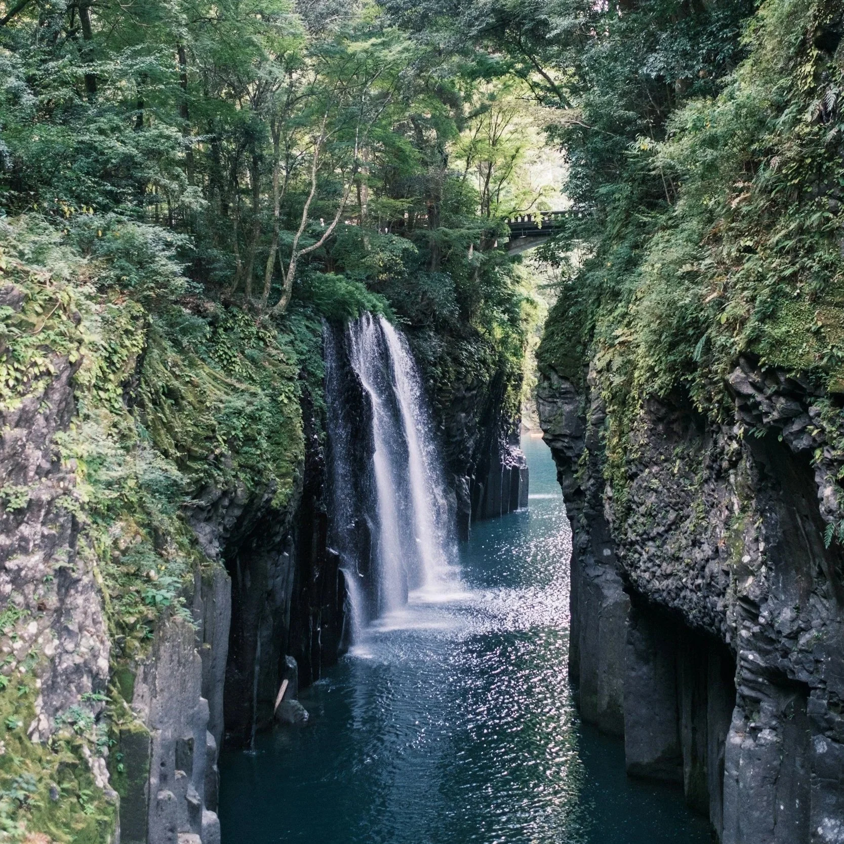 A narrow gorge with clear waters, with a red bridge crossing above the gorge and waterfalls streaming down the side of the gorge walls into the river