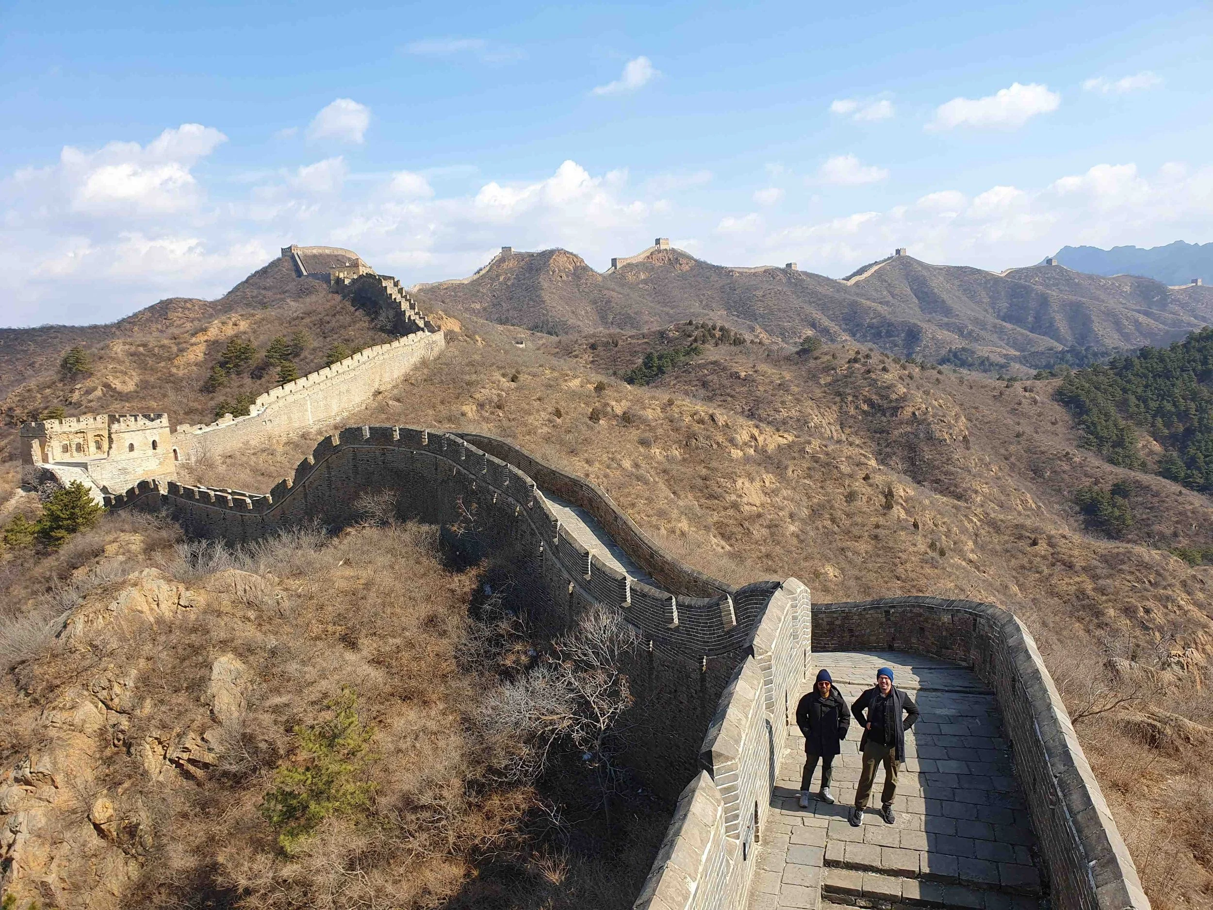 Two visitors standing on Jinshanling Great Wall in winter, the wall stretching across mountain ridgelines with multiple watchtowers visible into the distance