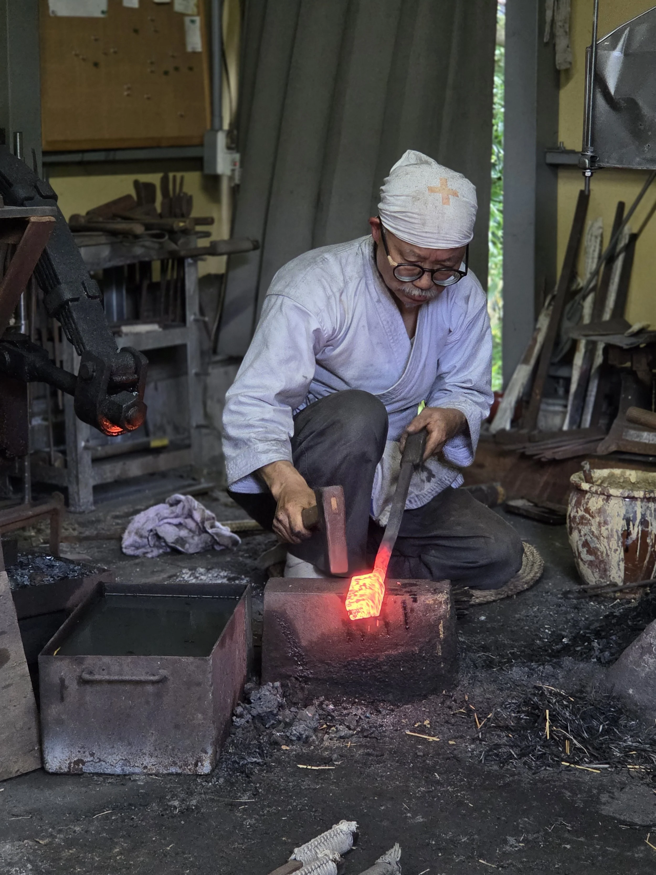 A blacksmith wearing glasses, a white headscarf with an orange cross, and a white coat working in a workshop. He is using a hammer on a red-hot piece of metal clamped in a vise. The workshop contains tools on shelves and a pegboard on the wall, with a dirt floor and various objects scattered around.