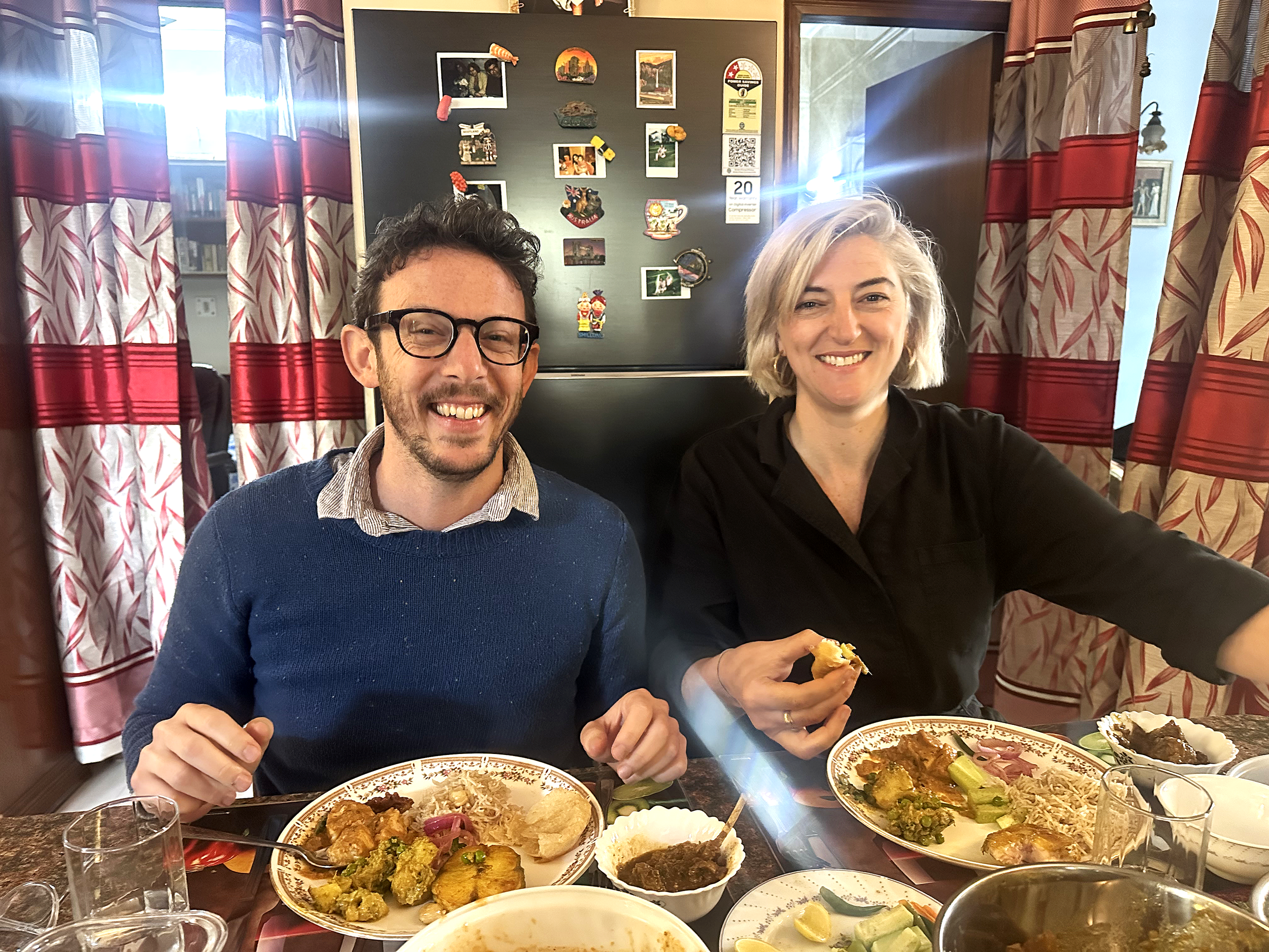 A man and a woman sit at a dinner table with plates of food, smiling at the camera in a cozy dining room with patterned curtains and a black board with pictures in the background.