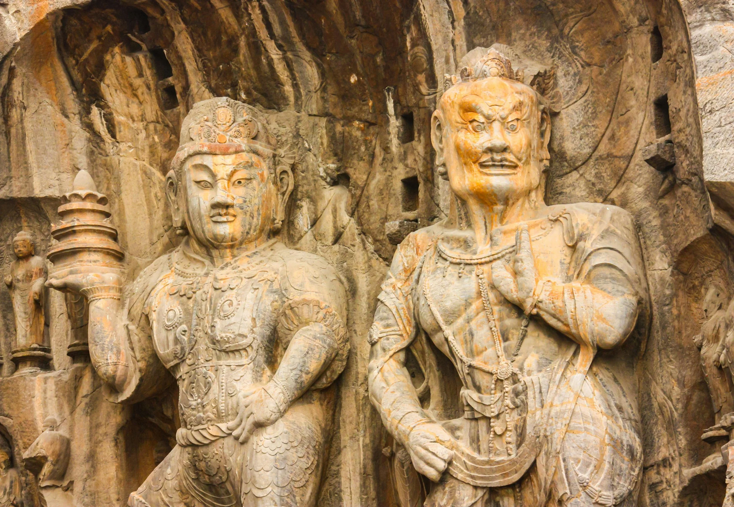 Carved stone statues of two warriors with intricate jewelry and armor, set against a rocky background, at the Longmen Grottoes in Longyang, Henan, China.