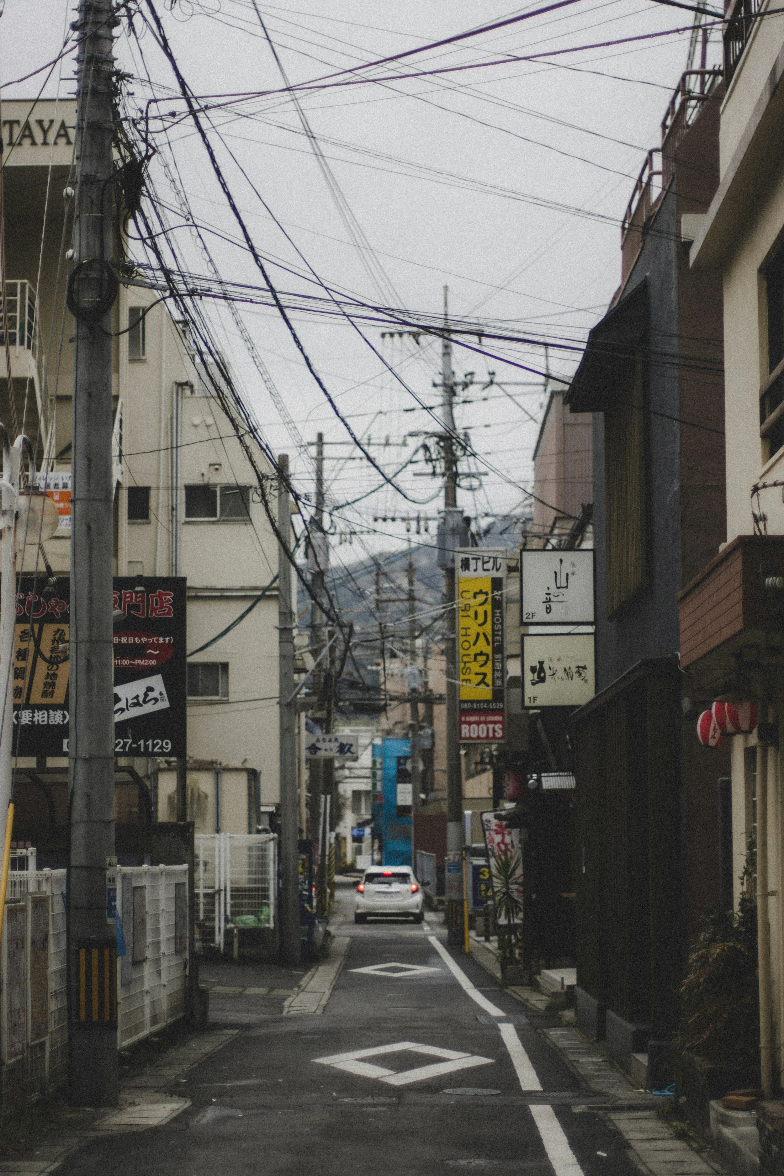 A car driving down a small one-way road in Japan, surrounded by shops and signs