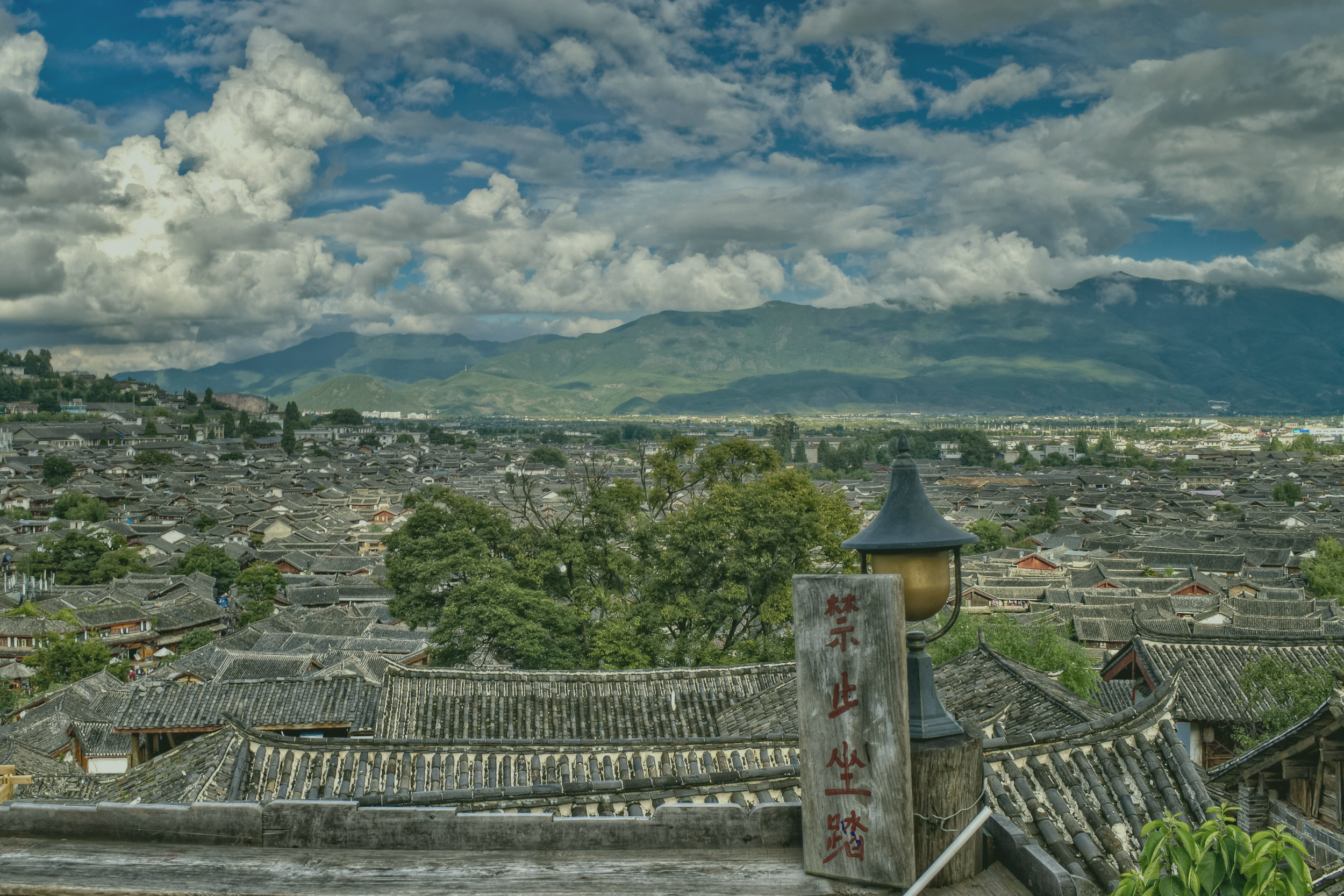 View of Lijiang, Yunnan, a UNESCO world heritage town in China with tiled rooftops, green trees, and mountains in the background under a cloudy sky. A wooden sign with red and black Chinese characters and a traditional lamp are in the foreground.