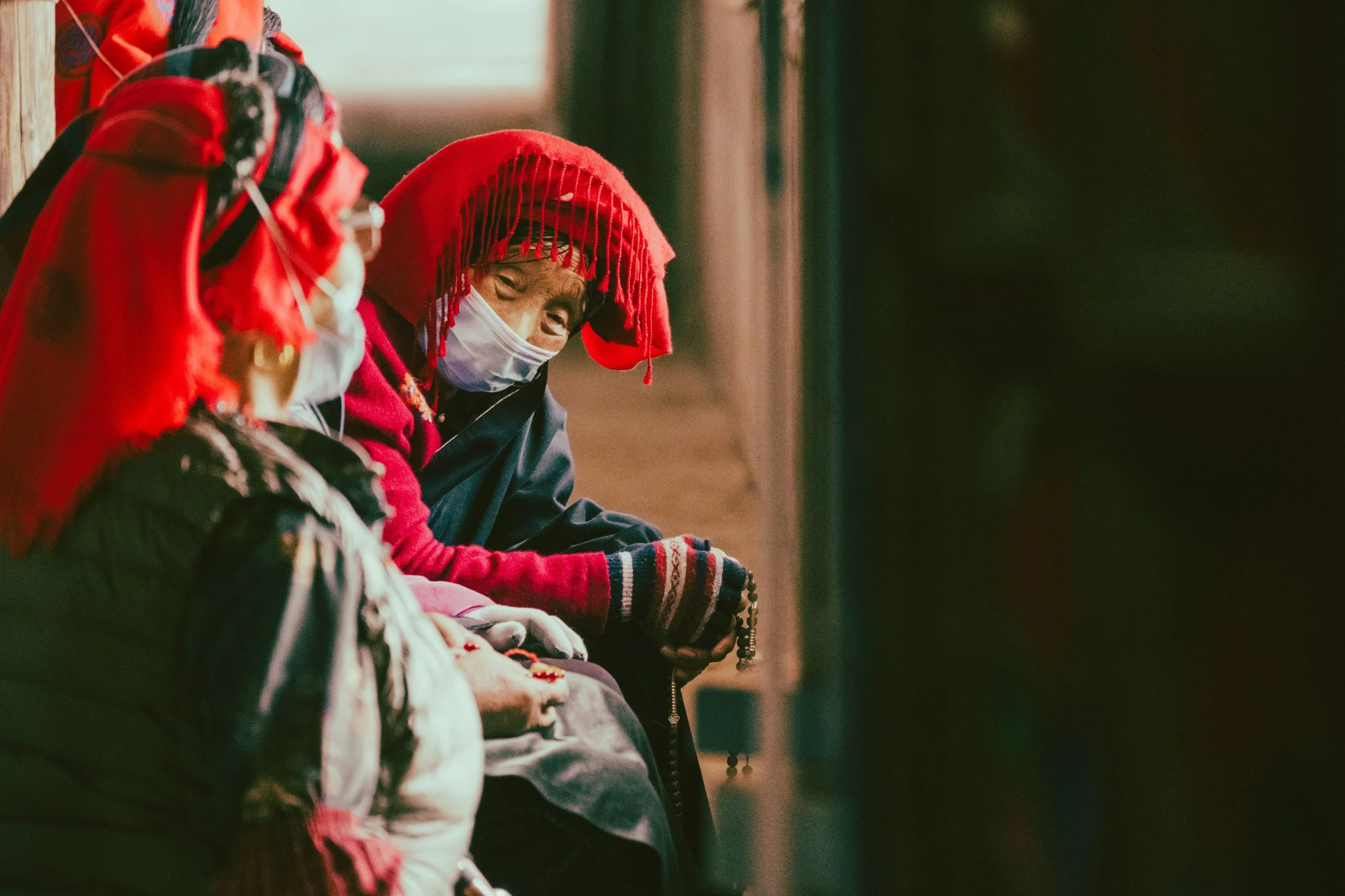 Tibetan ethnic minority people sitting on a bench, wearing traditional clothes and masks, with one person wearing a red hat and a scarf, another in a red sweater and knit gloves, in a cozy indoor setting.