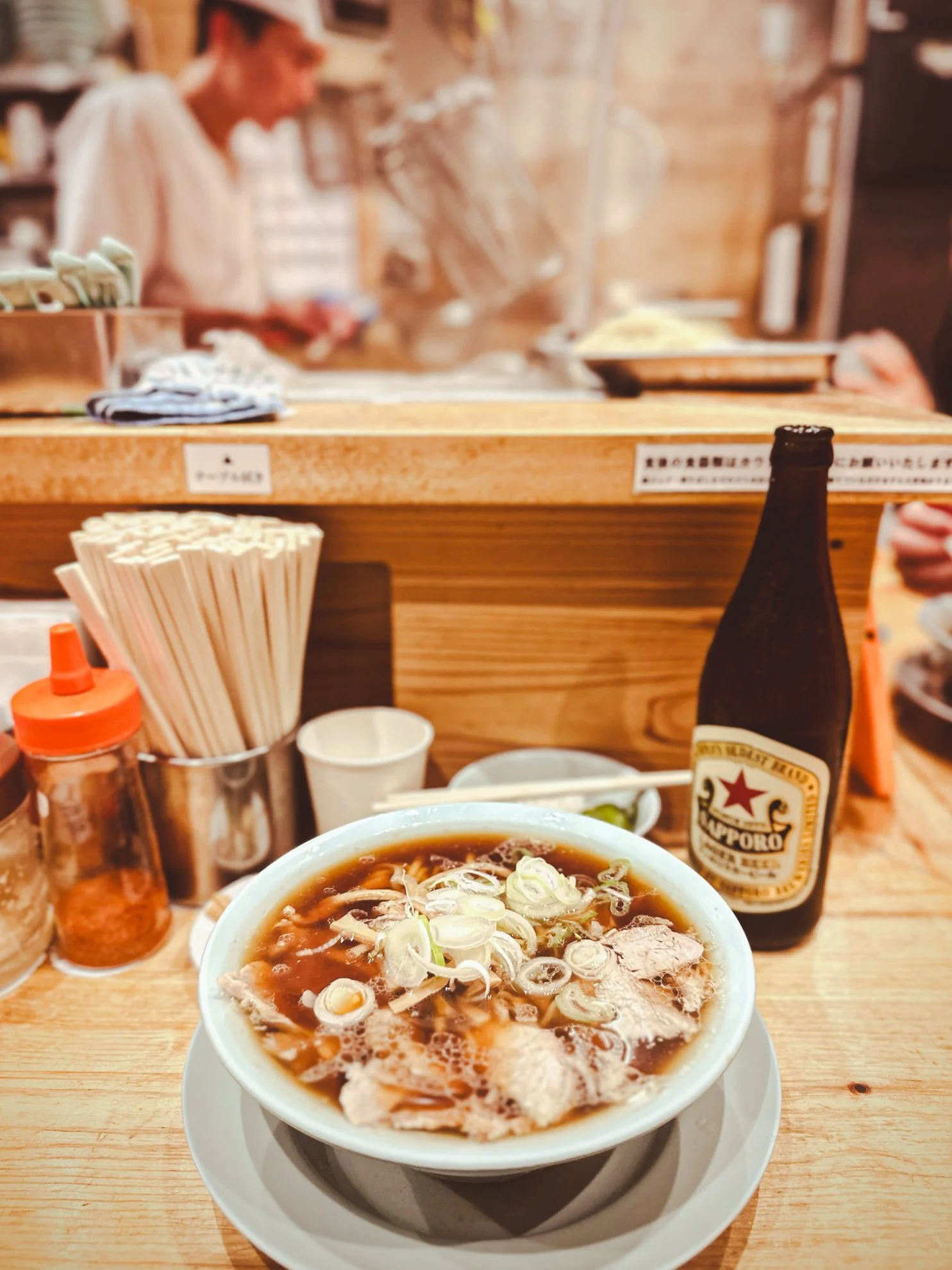 A bowl of ramen and a sapporo beer at a bar counter in Shimokitazawa, Tokyo, with a chef in the background preparing food