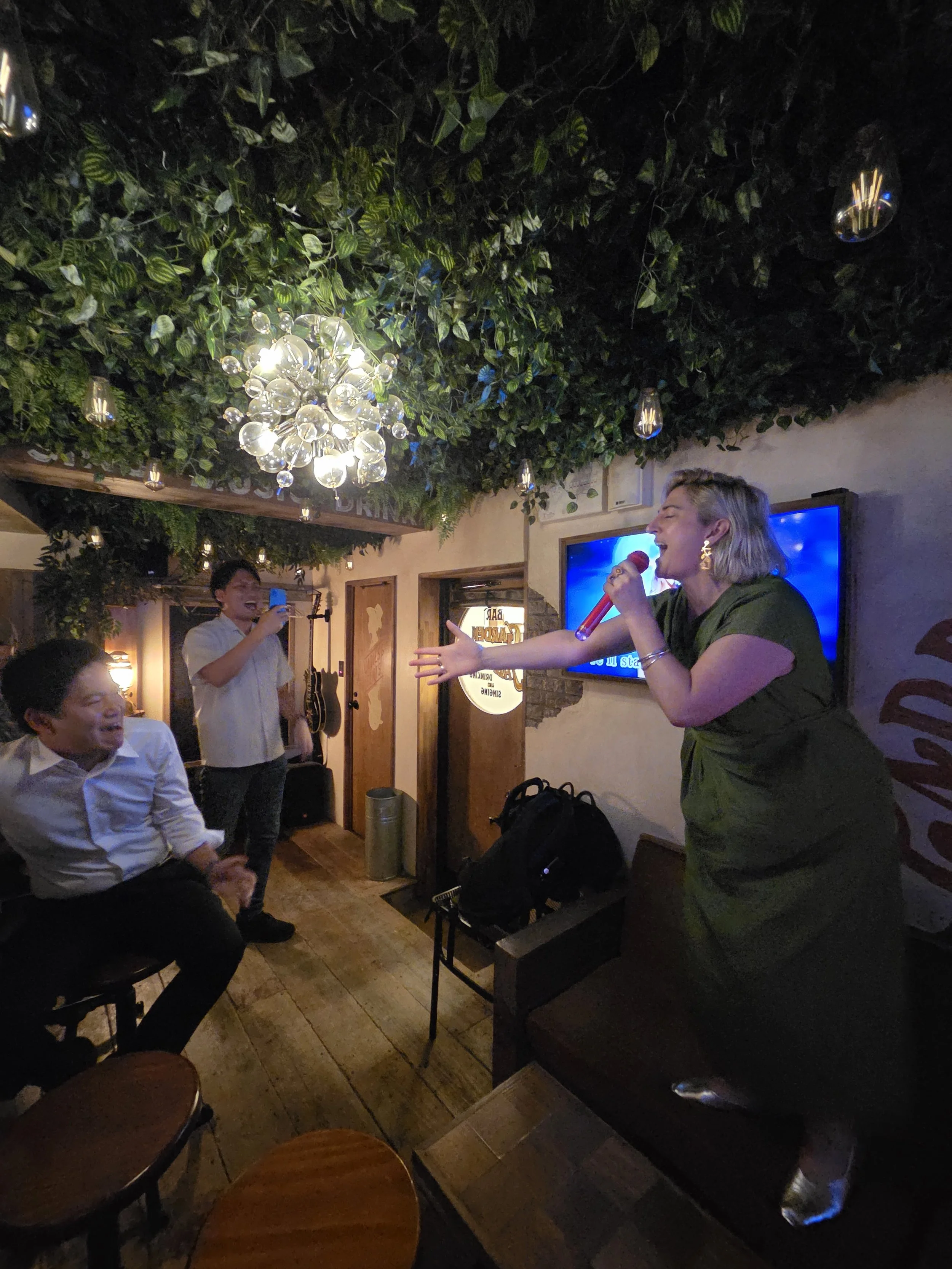 A woman in a green dress singing karaoke with two Japanese salarymen in a bar decorated like a camping site in Shimokitazawa, Tokyo