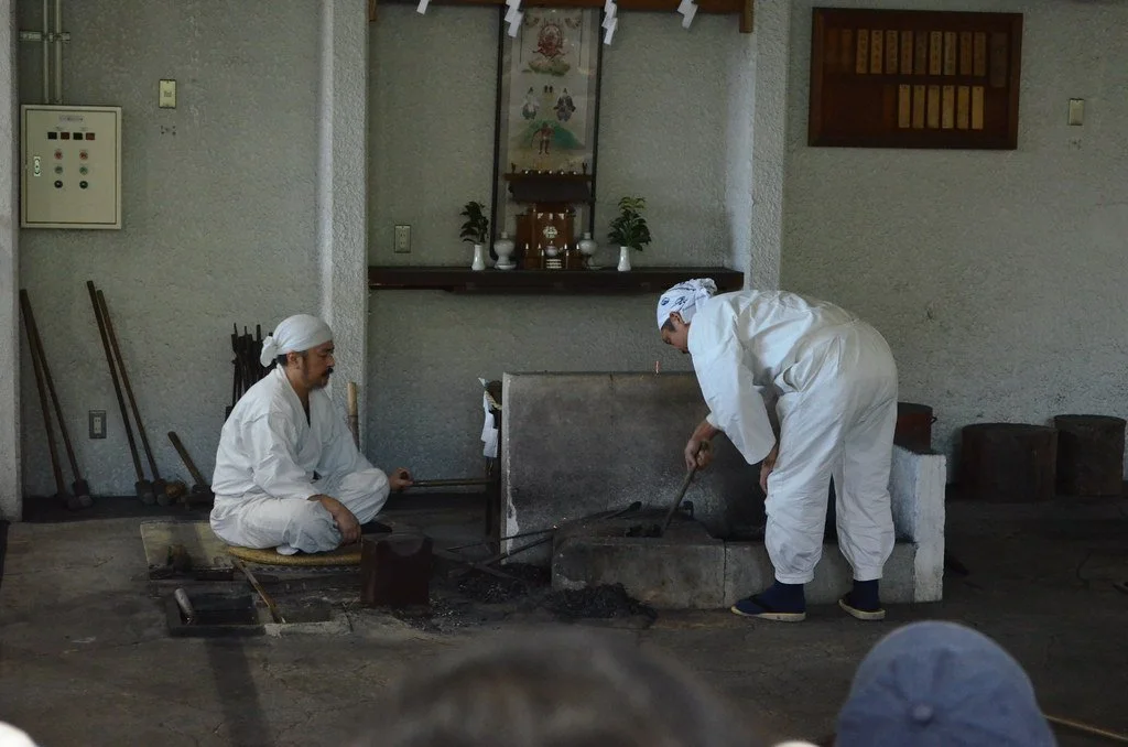 Two swordsmiths in traditional white clothing working at a forge during a demonstration at the Seki Swordsmith Museum, Gifu