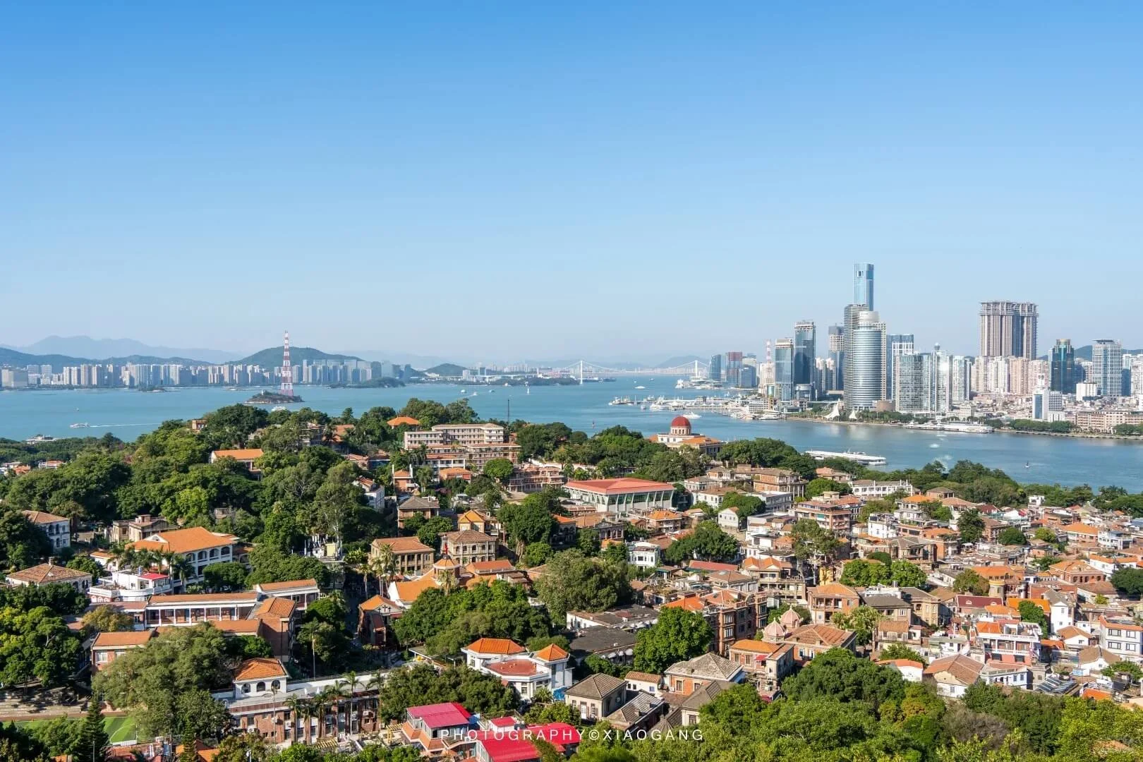 A cityscape of Xiamen featuring the river separating the modern high-rise buildings and skyscrapers of the city, and a quieter residential area on Gulangyu with houses and trees on the west side, under a clear blue sky.