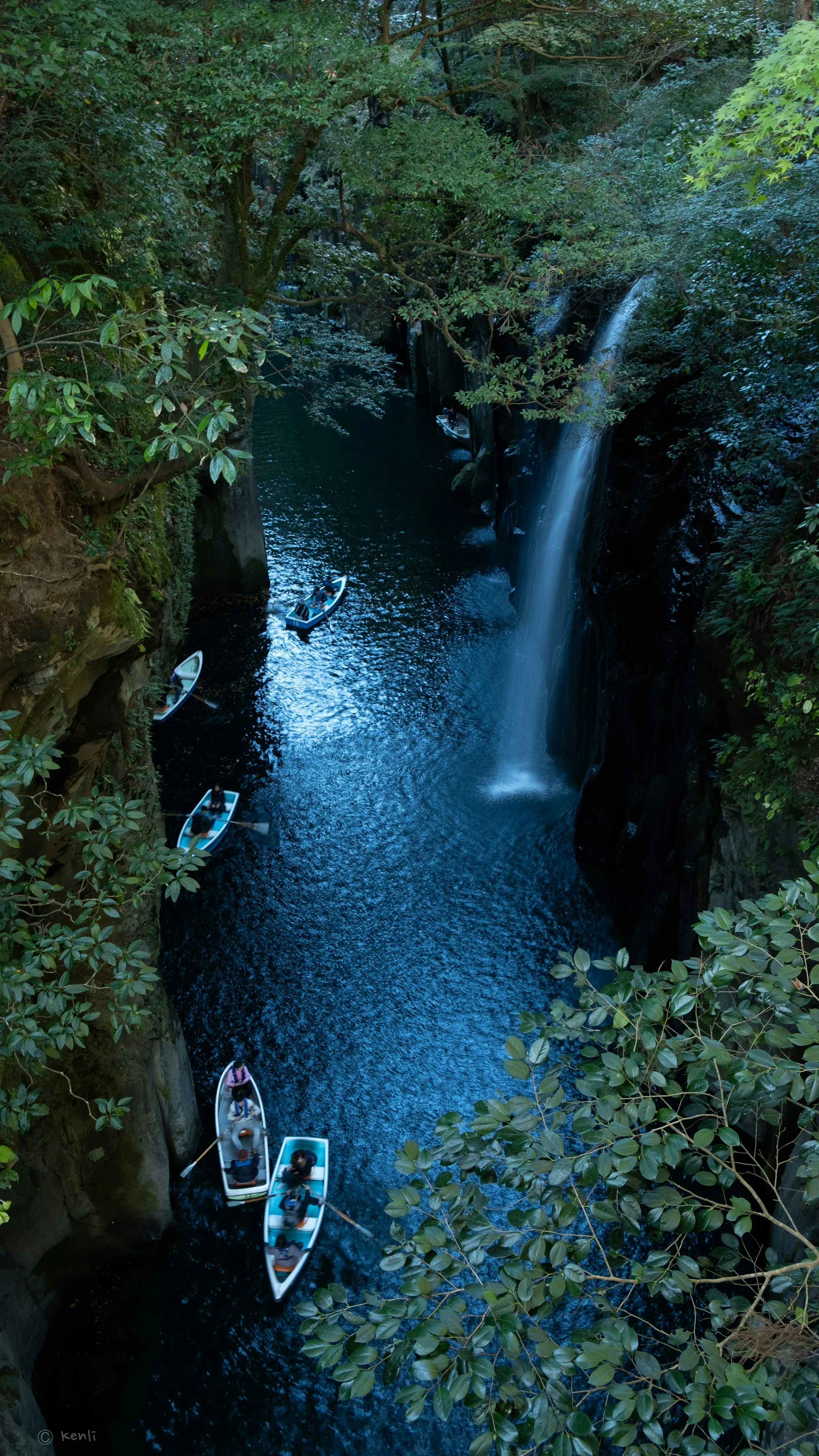 A narrow river with a waterfall on the right side, surrounded by lush green trees in Takachiho Gorge, Miyazaki, Japan. Several small boats with people are floating on the water, heading towards the waterfall.