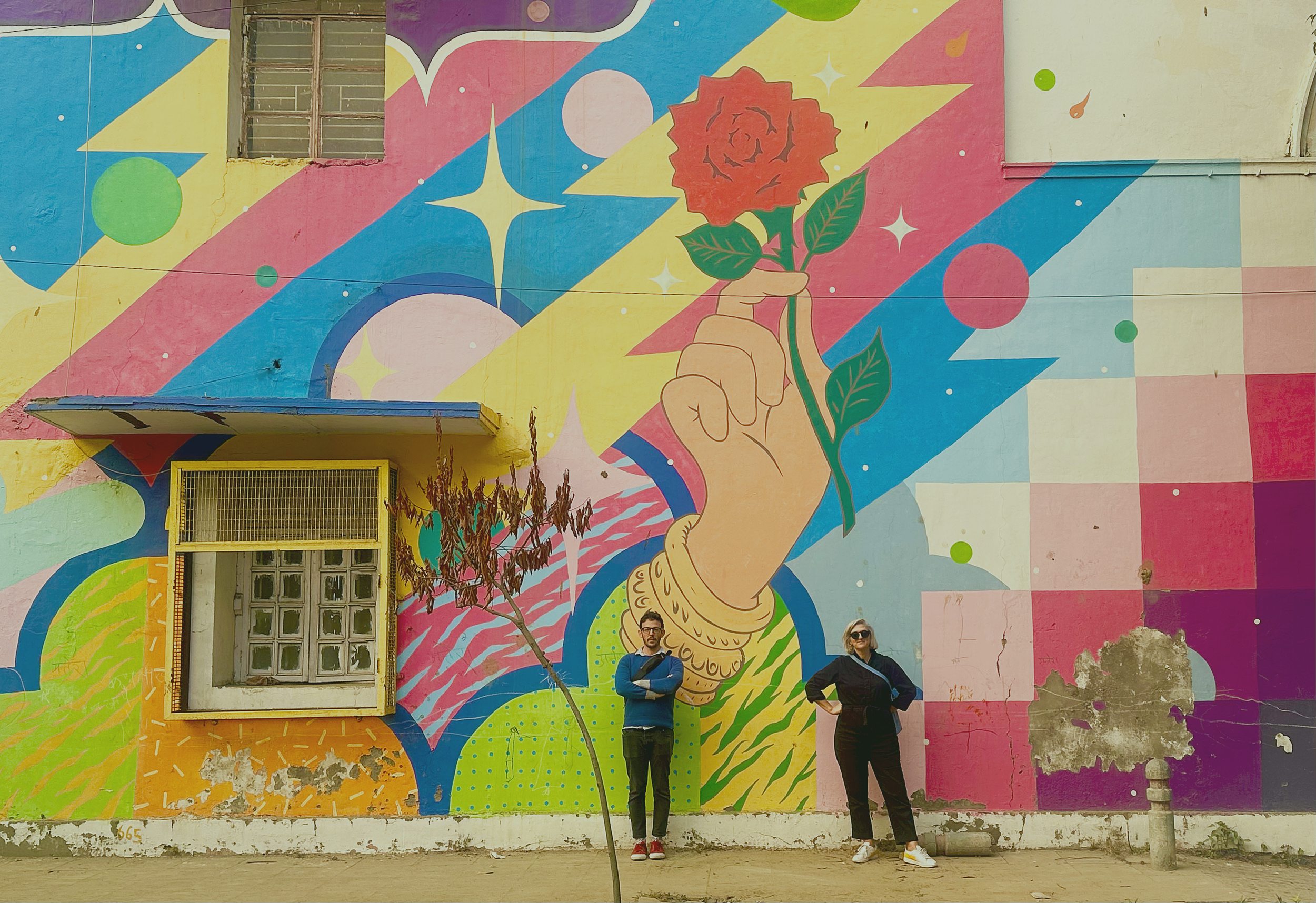 A colorful mural on a building wall featuring a large hand holding a rose. Two people stand in front of the mural, one man with glasses and a woman with sunglasses, both striking confident poses.