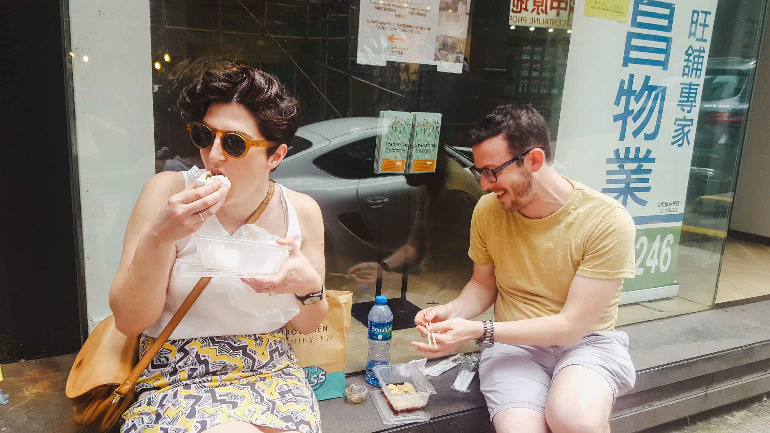 A woman wearing sunglasses and a patterned skirt is dim sum from a plastic container while sitting next to a smiling man in a yellow shirt and shorts. They are sitting outside near a store window with signs in Hong Kong.