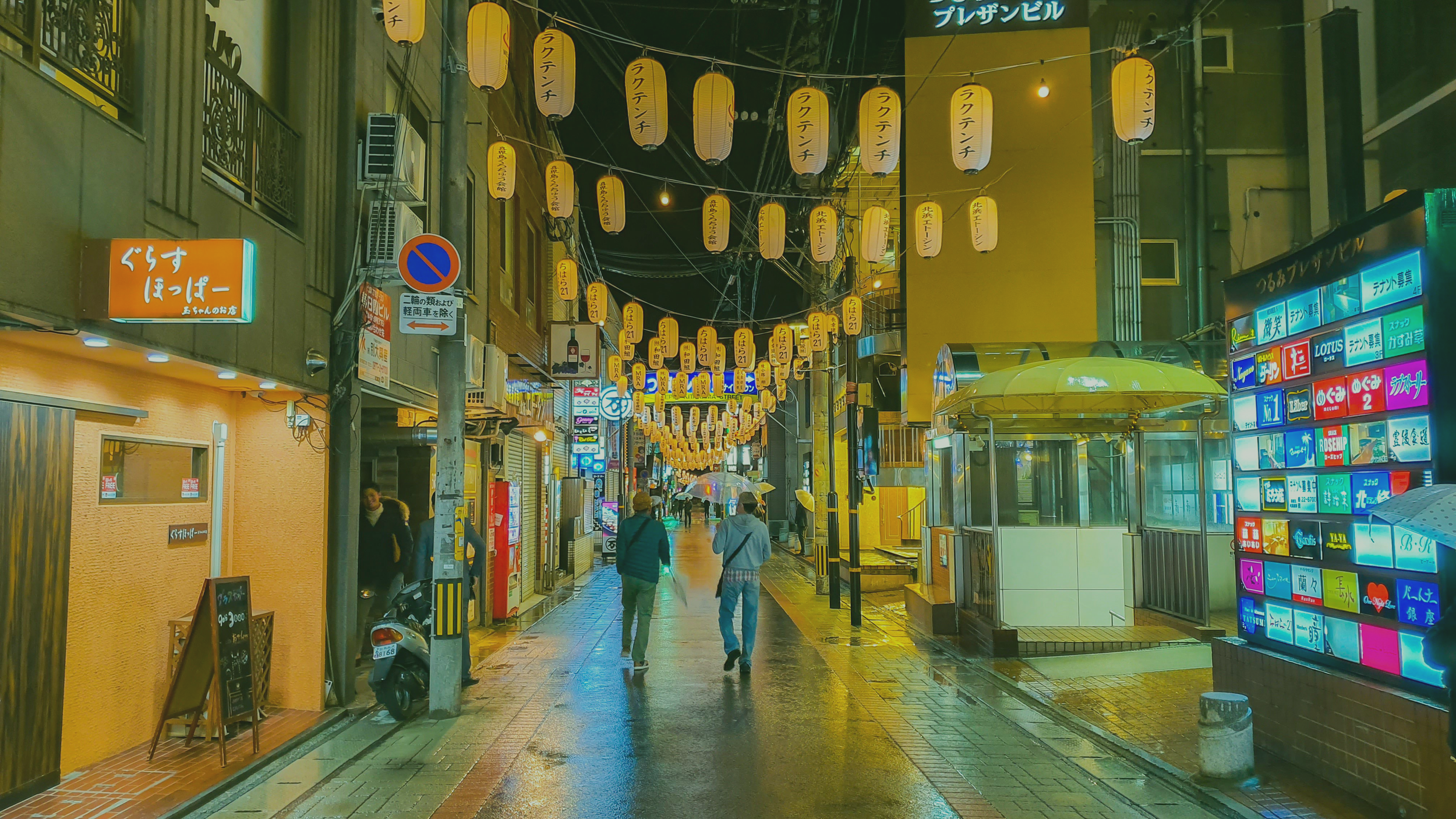 Night view of a decorated street in Beppu, Oita, Japan with yellow lanterns hanging overhead, wet pavement reflecting lights, people walking with umbrellas, illuminated signs and storefronts along the street.