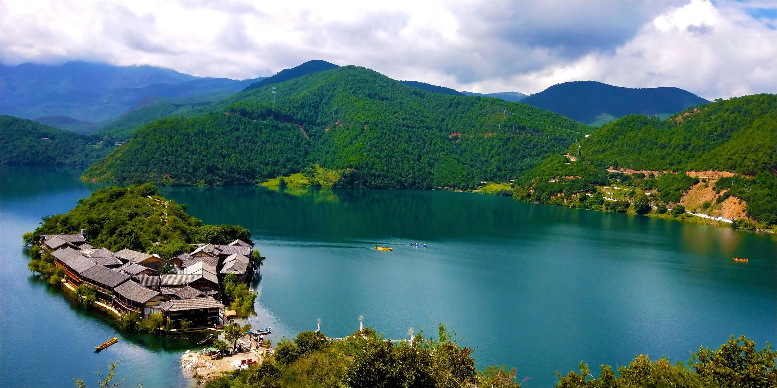 A scenic view of large, calm Lugu Lake in Yunnan, China surrounded by green mountains under a cloudy sky. There is a small island with buildings and trees in the foreground, and boats on the water.