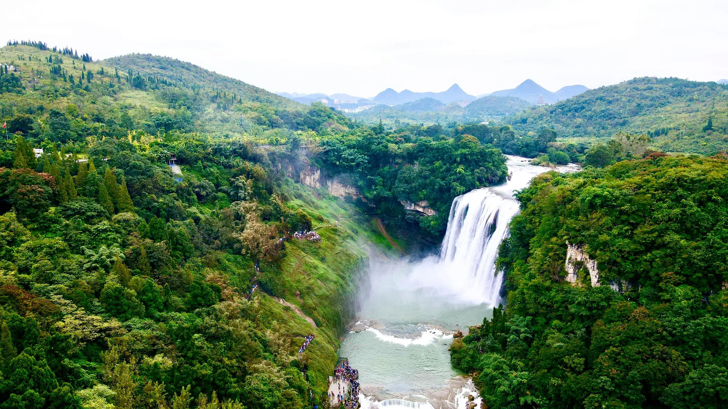 Lush green landscape in Guizhou, China with a large waterfall cascading into a river, surrounded by dense forested hills and mountains in the background.