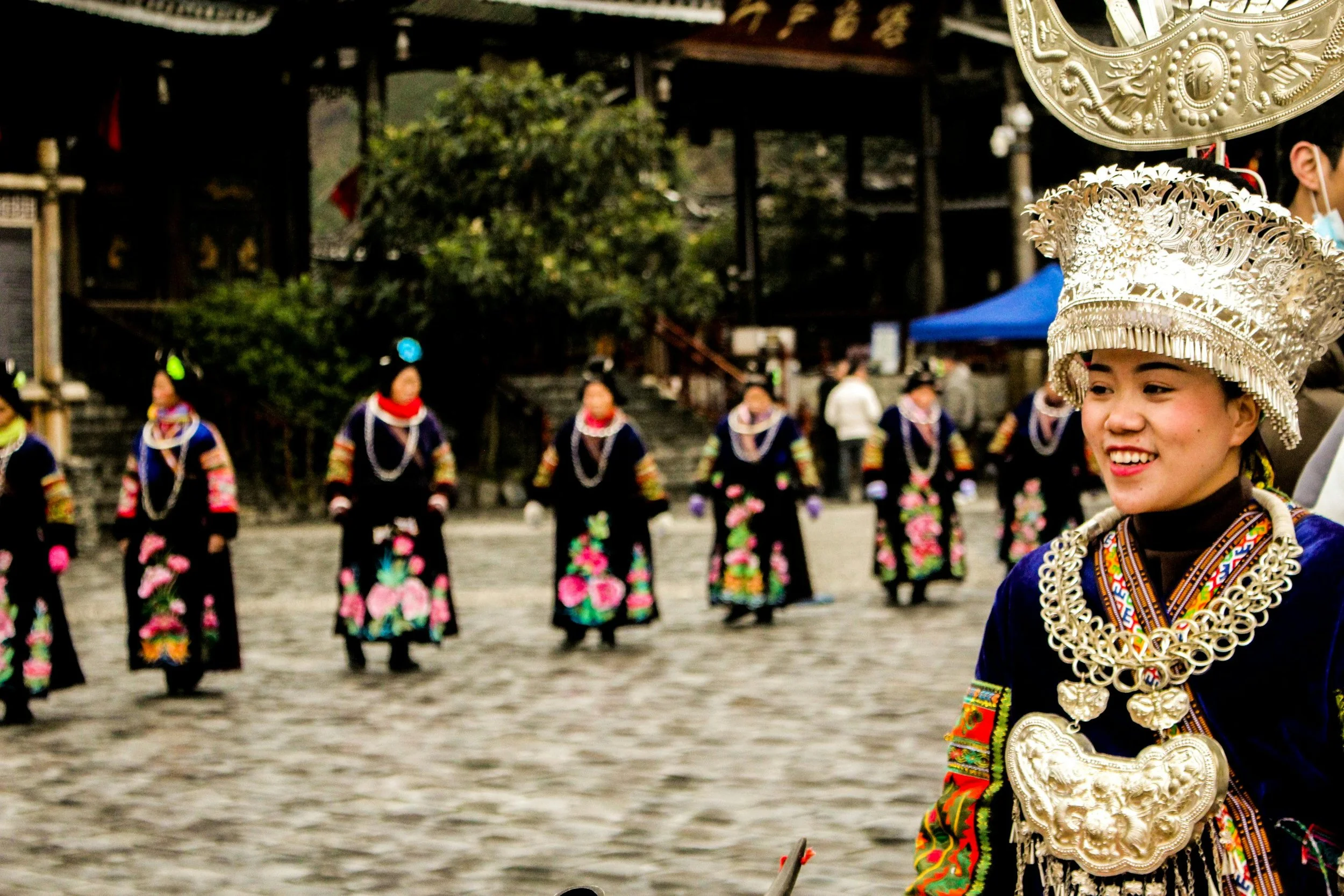 Miao minority women dressed in traditional colorful attire and elaborate silver jewelry, participating in a cultural dance in an outdoor setting in Guizhou, China.