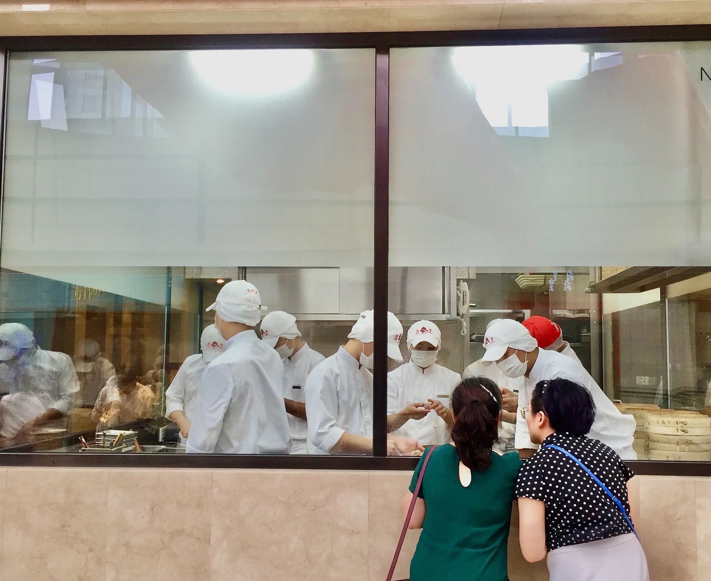 Two women looking through a window into the kitchen at Din Tai Fung in Shanghai, watching chefs make xiaolongbao soup dumplings