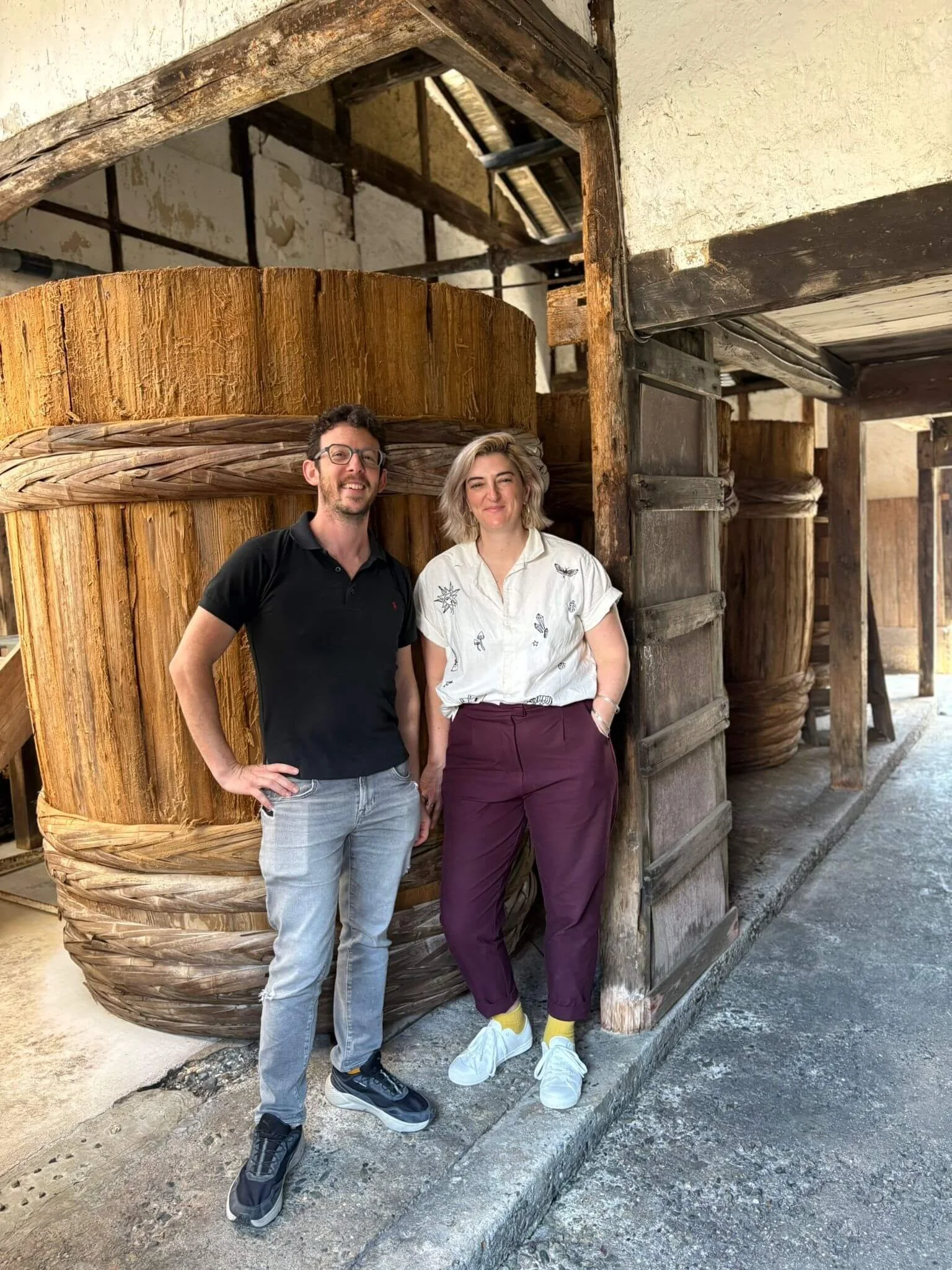 Minsky and Beth standing next to each other outdoors in front of large wooden barrel, full of aging soy sauce, in a rustic wooden structures.