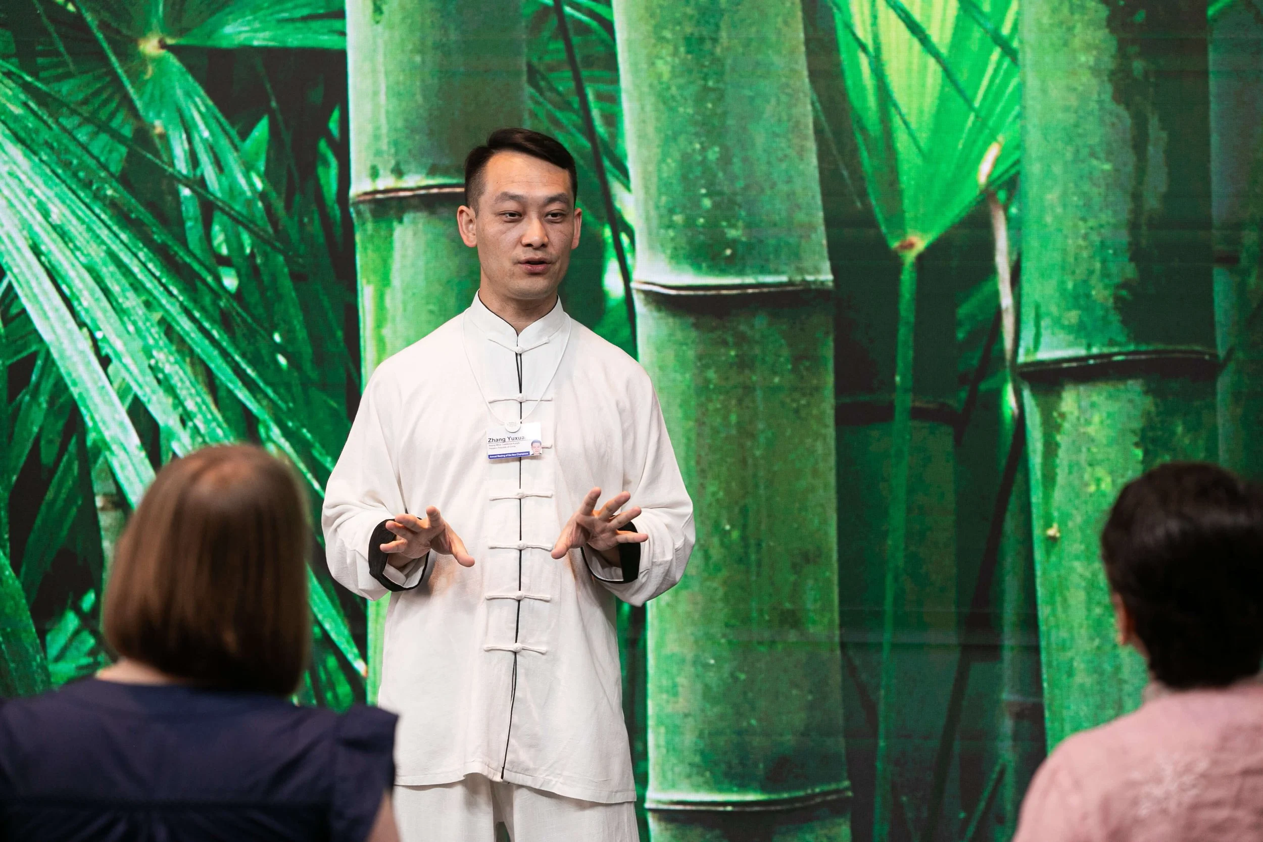 A man, Yuxuan Zhang, in traditional white Chinese attire giving a presentation about tai chi to two women in front of a large, vibrant green bamboo background.