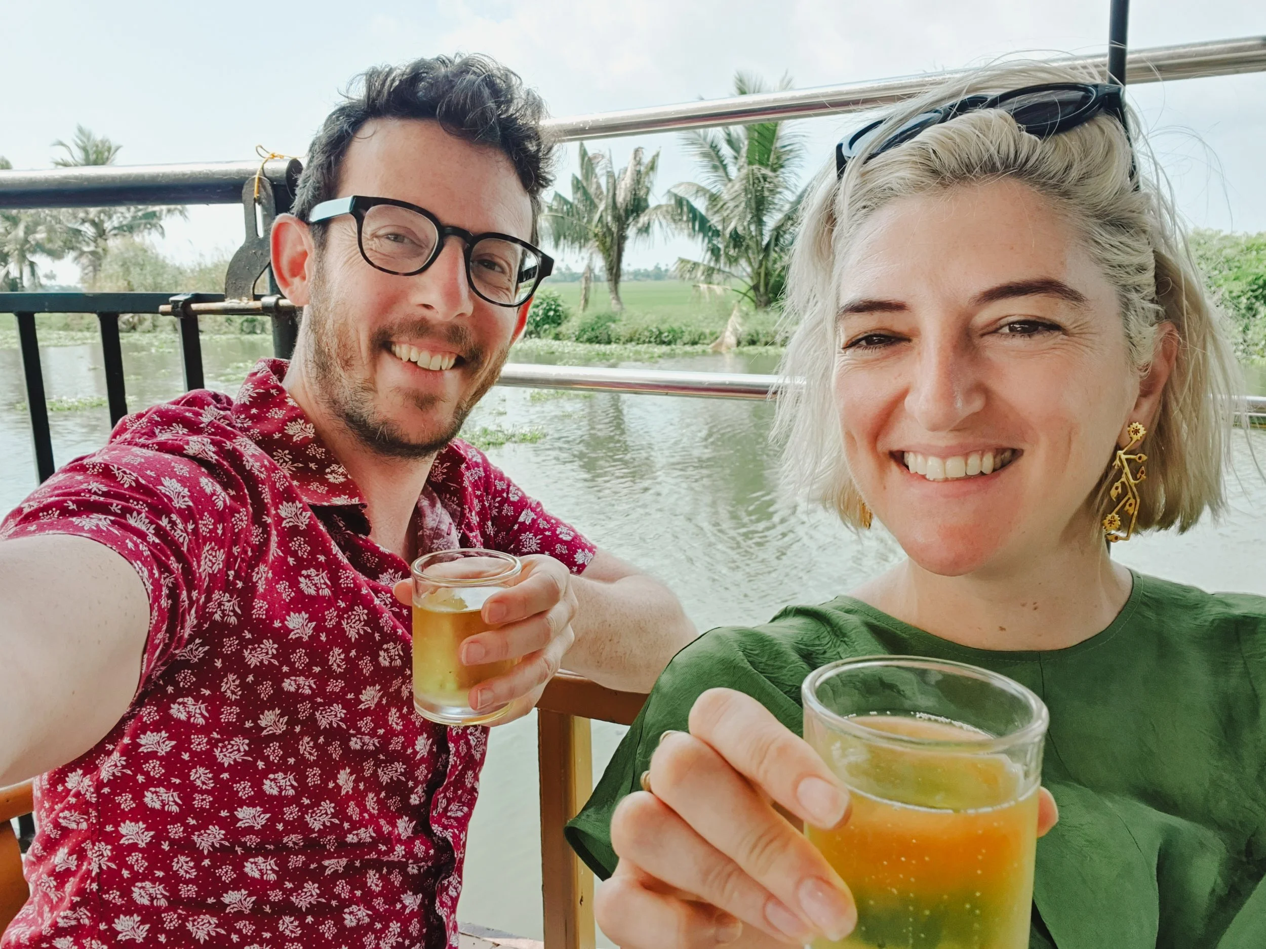 A smiling man and woman holding drinks while taking a selfie outdoors by a river with palm trees and greenery in the background.