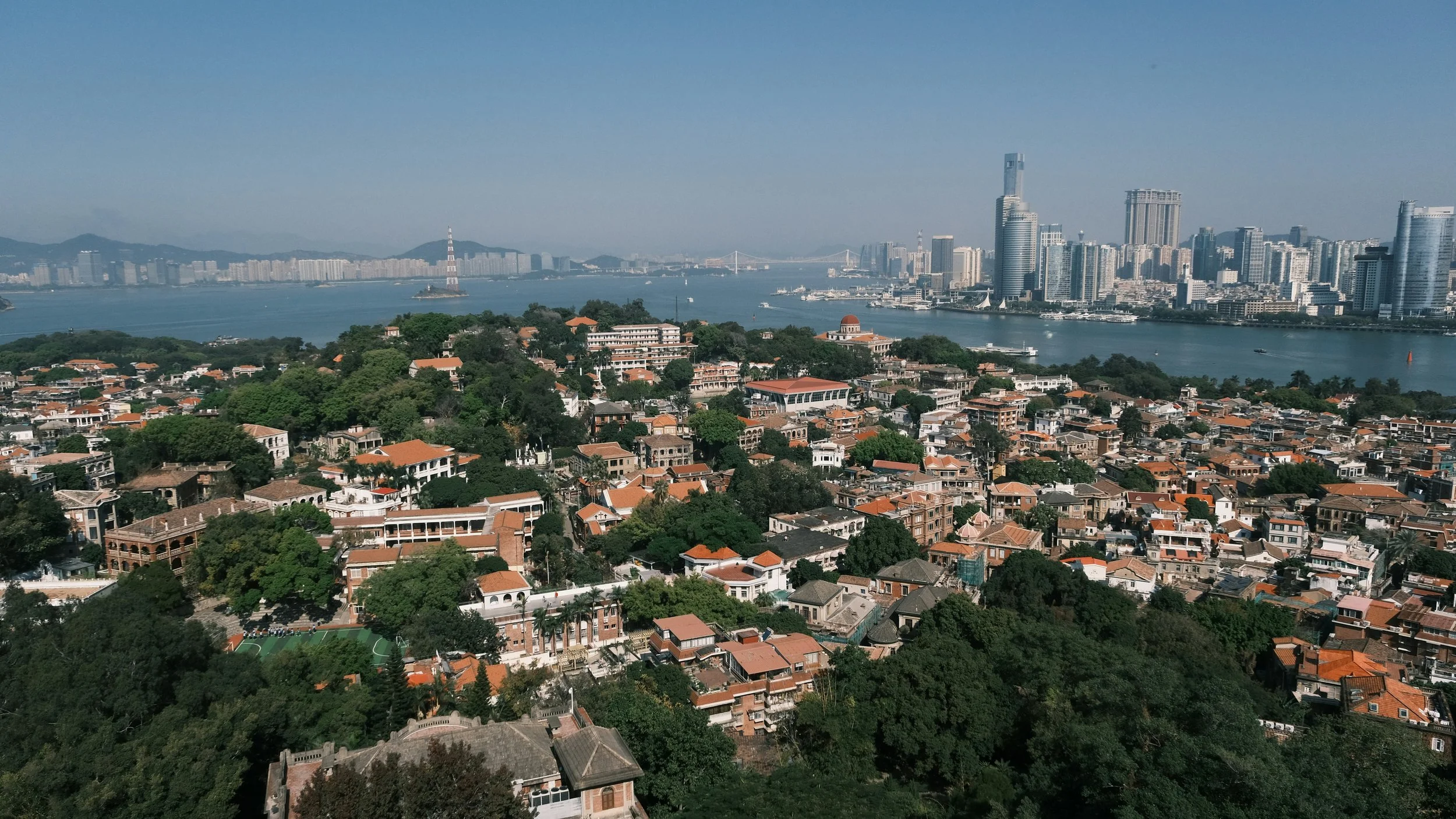A cityscape in Xiamen with the ocean in the foreground, numerous residential buildings with orange roofs on gulangyu, and a skyline of tall modern buildings and skyscrapers in the background.