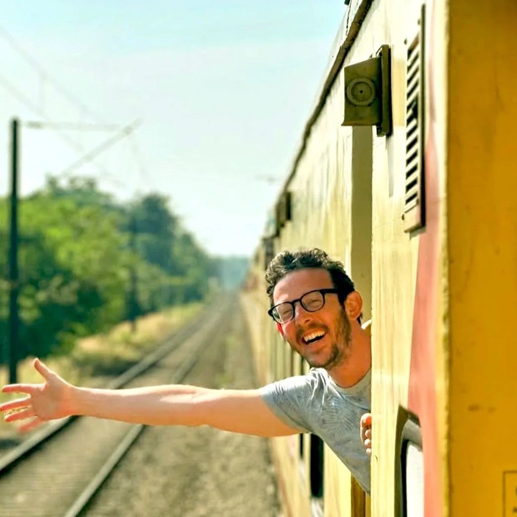 A man with glasses and a beard leaning out of a train window, smiling and extending his arm with traffic in the background.