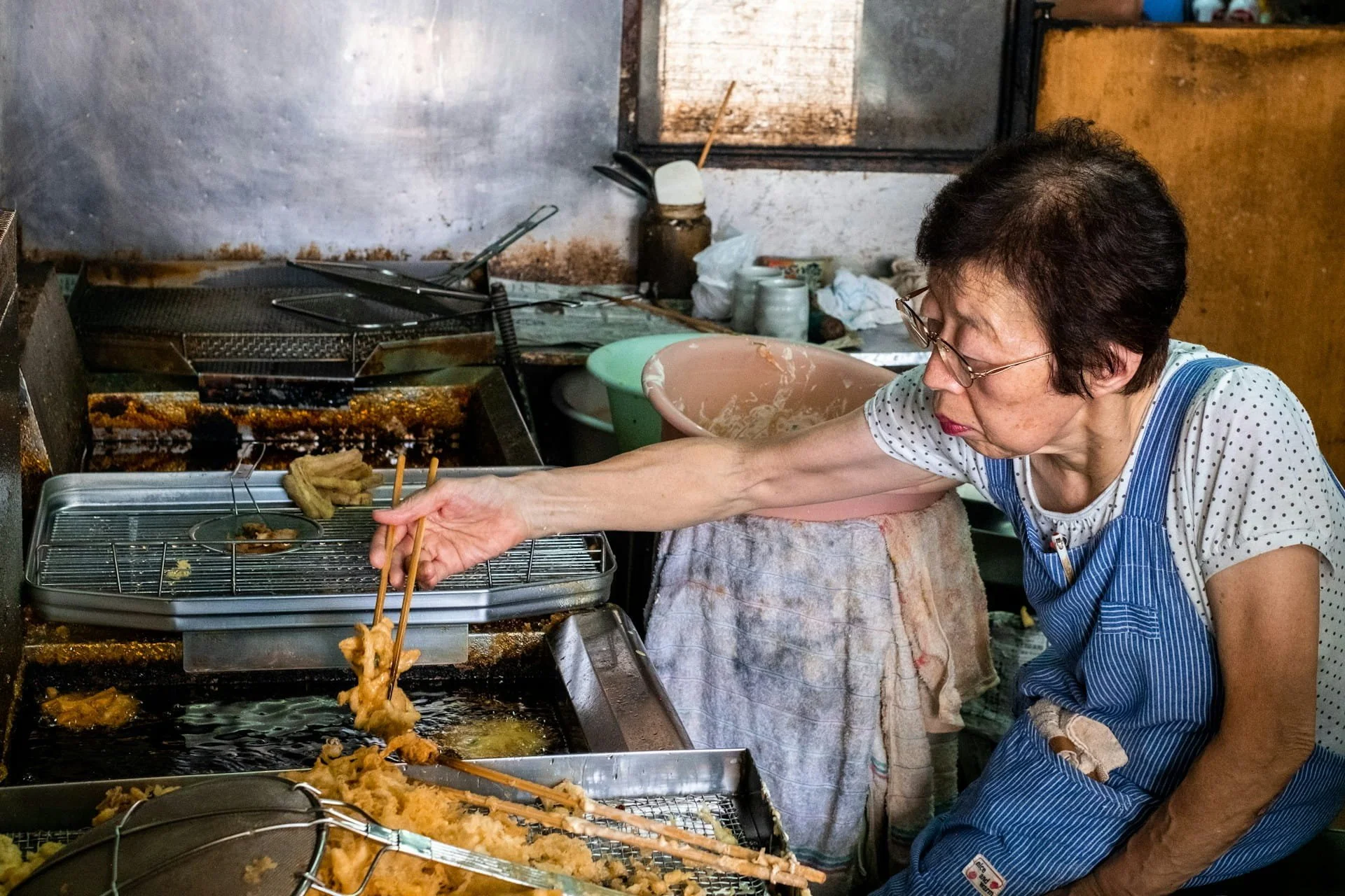 apanese woman frying tempura in a well-worn kitchen, chopsticks reaching into the oil