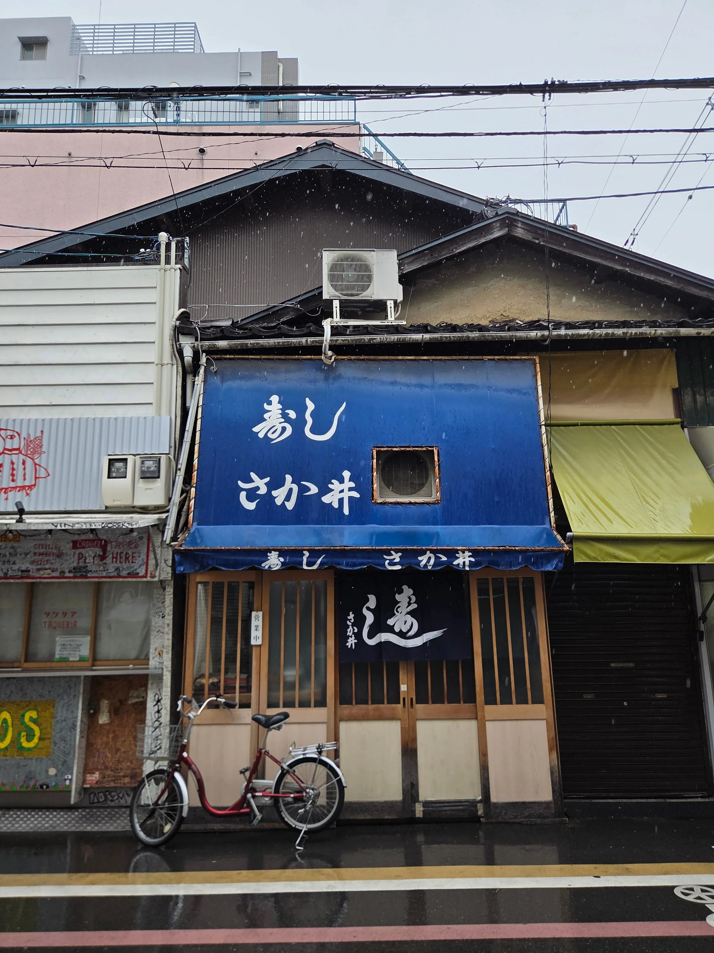 Exterior of Sakai sushi restaurant in Kyoto on a rainy day, with a faded blue awning and bicycle parked outside