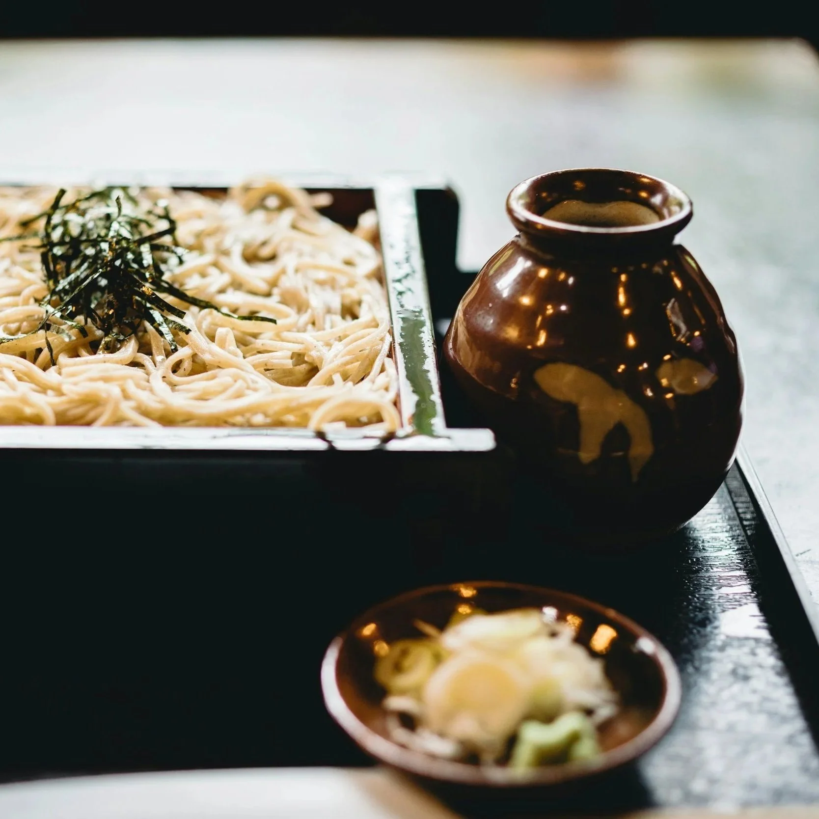 A tray of cold soba noodles garnished with shredded nori seaweed next to a small dark brown ceramic pitcher and a small dish of sliced green onions.