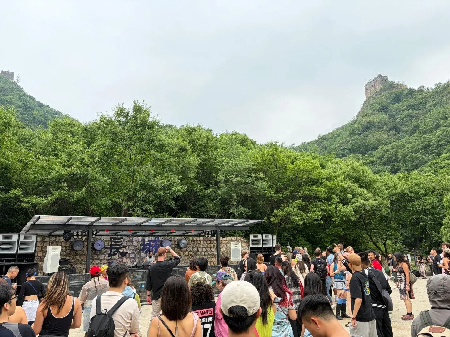Great Wall Festival crowd dancing in front of a DJ stage, with the Great Wall and watchtowers visible on the hillside behind the treeline, Huairou District Beijing