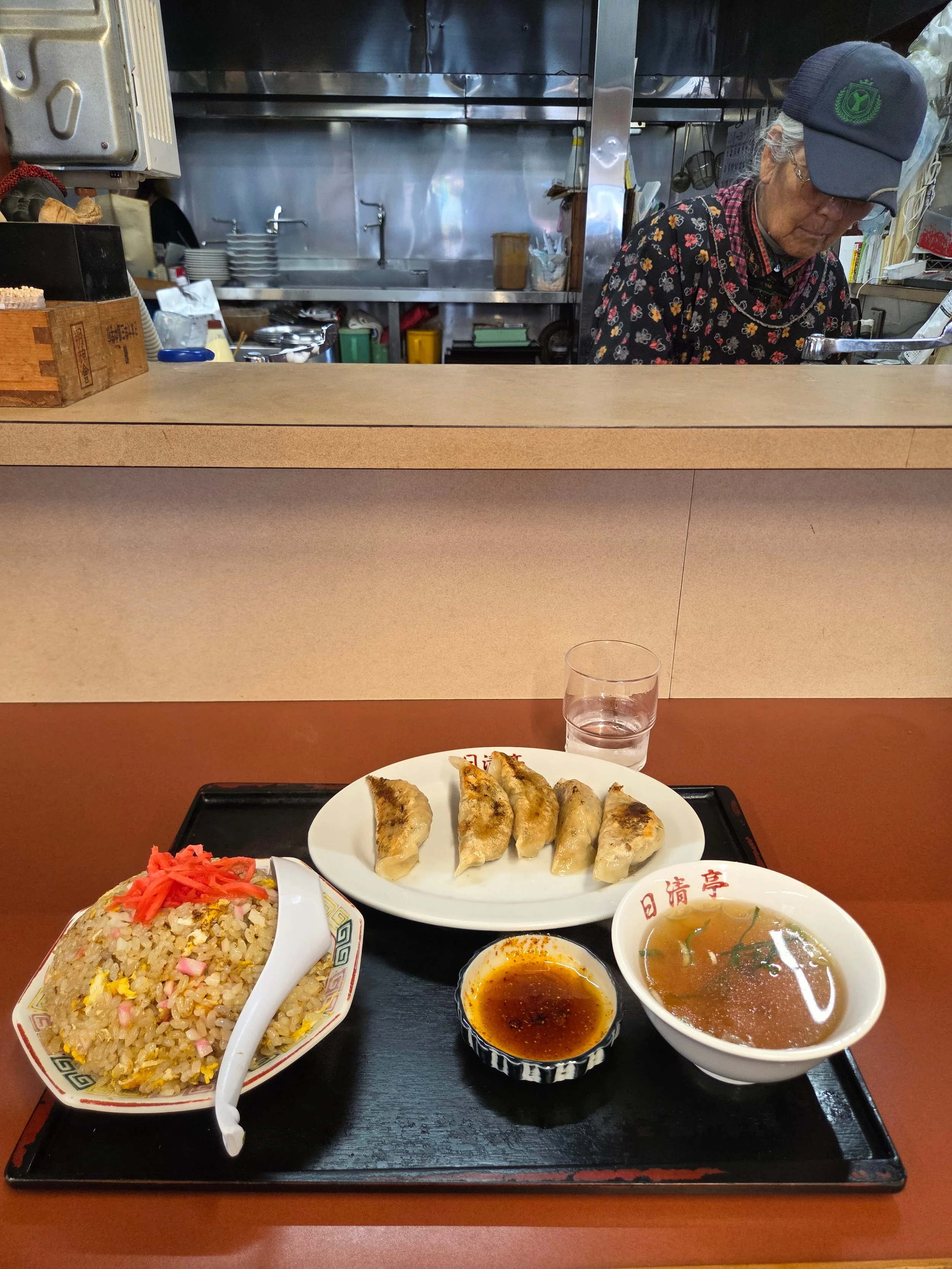 Gyoza, fried rice and soup set served at a small Japanese shokudo, with elderly woman cooking in the background