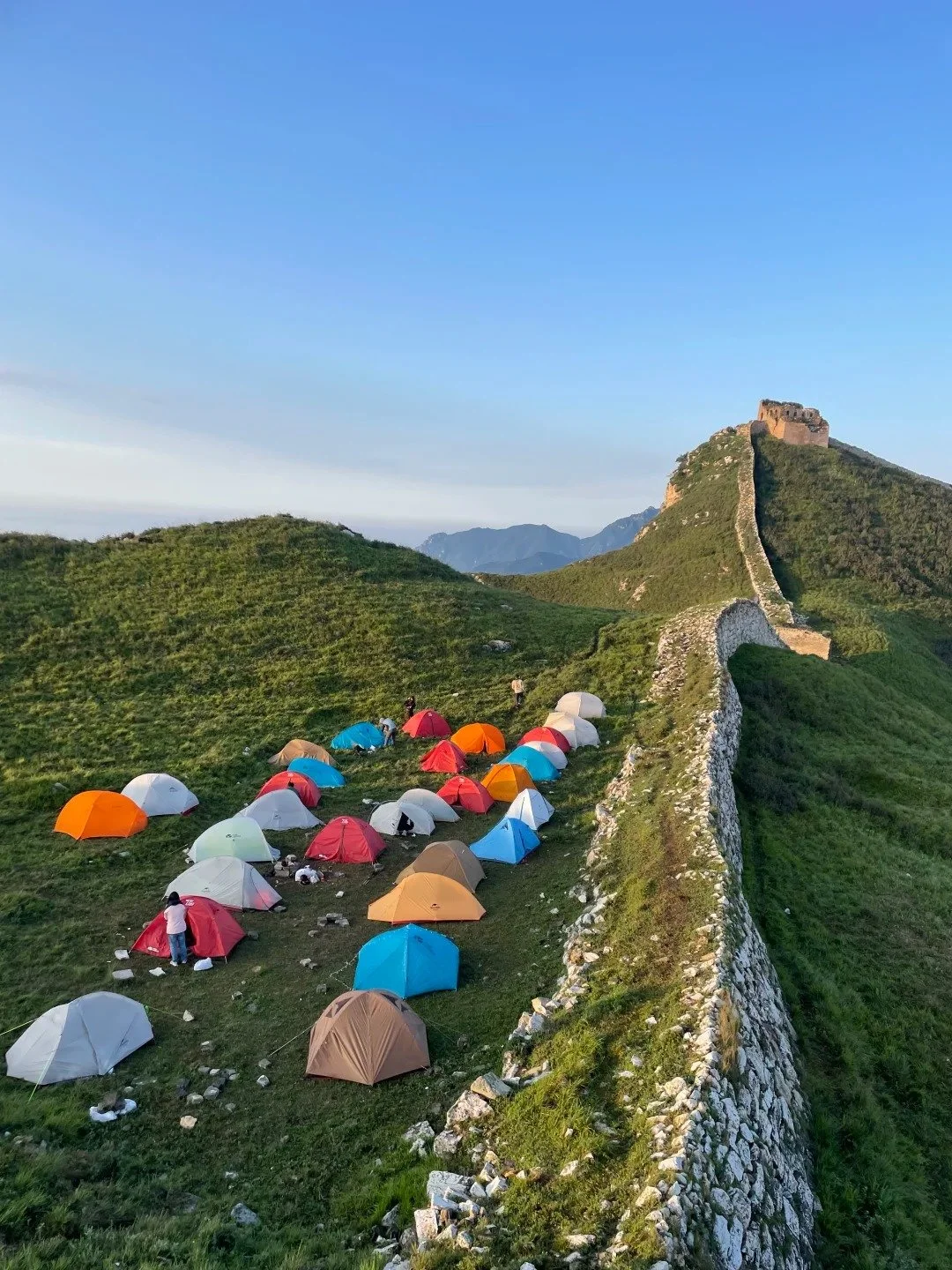 Colourful camping tents pitched alongside the Great Wall at dusk, the wall and a watchtower visible stretching up the hillside behind