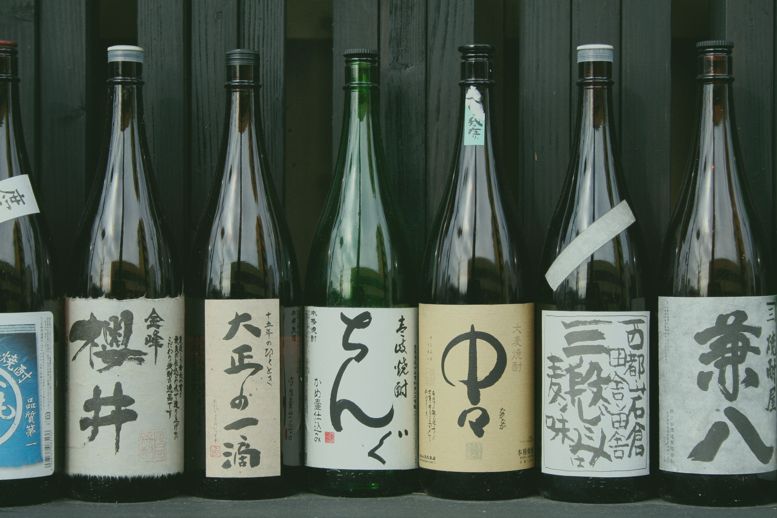 A row of seven bottles of sake with Japanese labels, arranged against a wooden backdrop.
