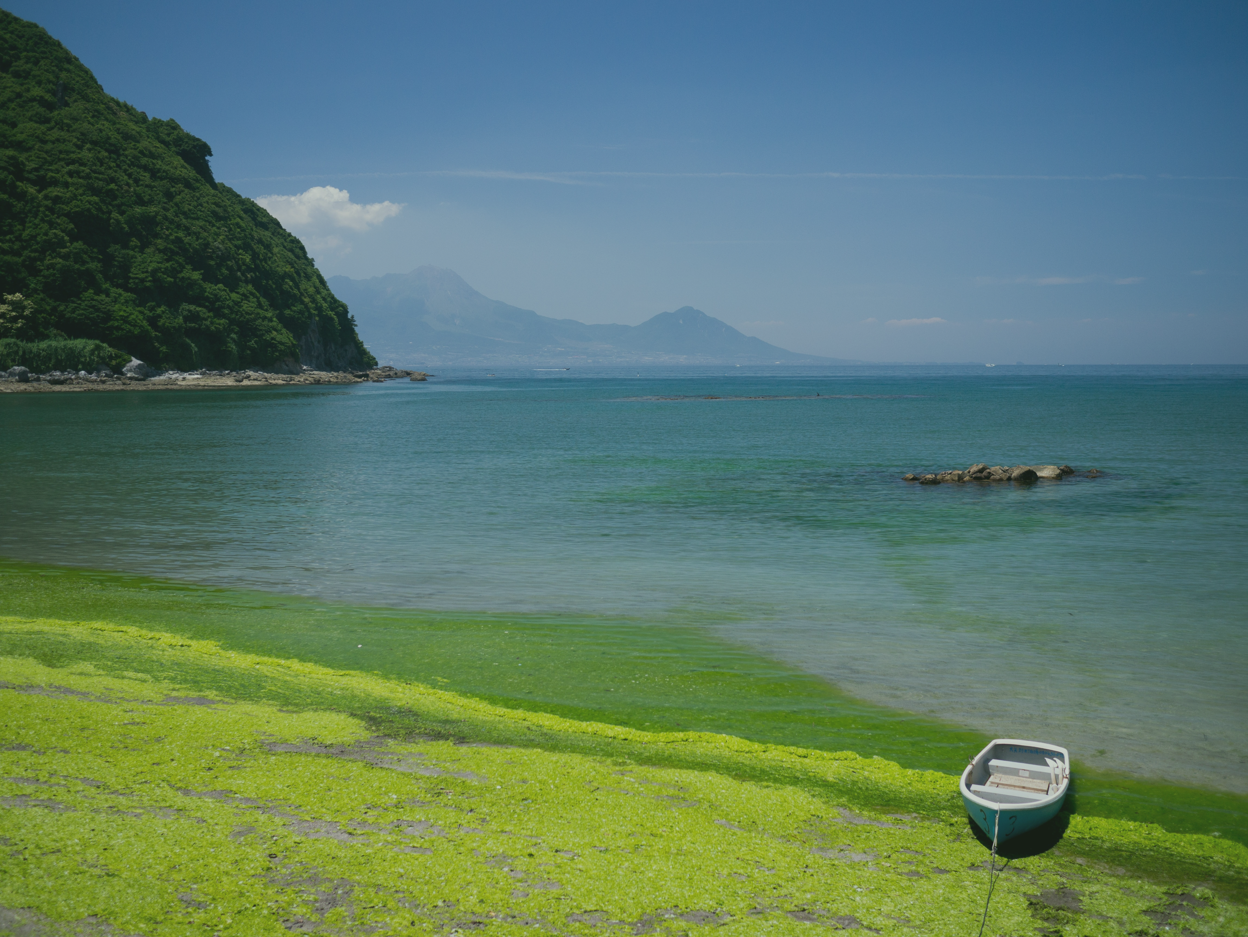 A small boat resting on a grassy, algae-covered shoreline with calm water in the Amakusa region of Kumamoto, Japan, a green hillside on the left, and mountains in the background under a blue sky.