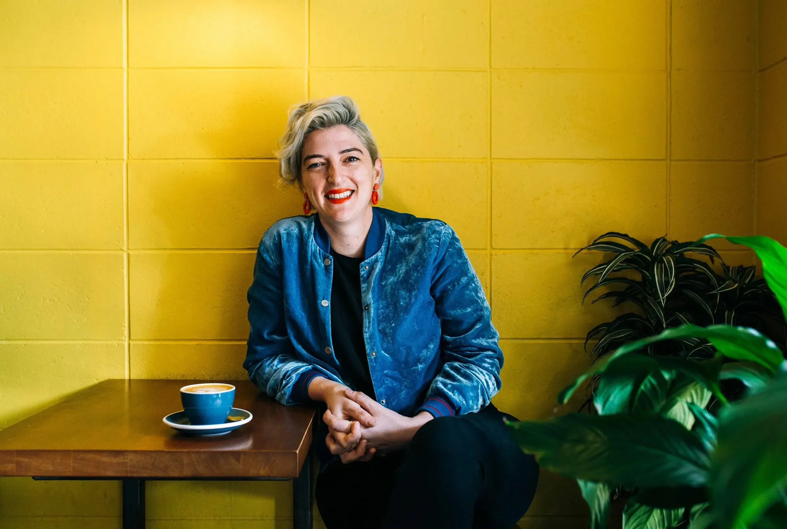 A woman with short wavy blonde hair, wearing a blue velvet jacket, red earrings, and red lipstick, sitting at a wooden table with a blue coffee cup and saucer, smiling in front of a yellow tiled wall with green plants on the side.
