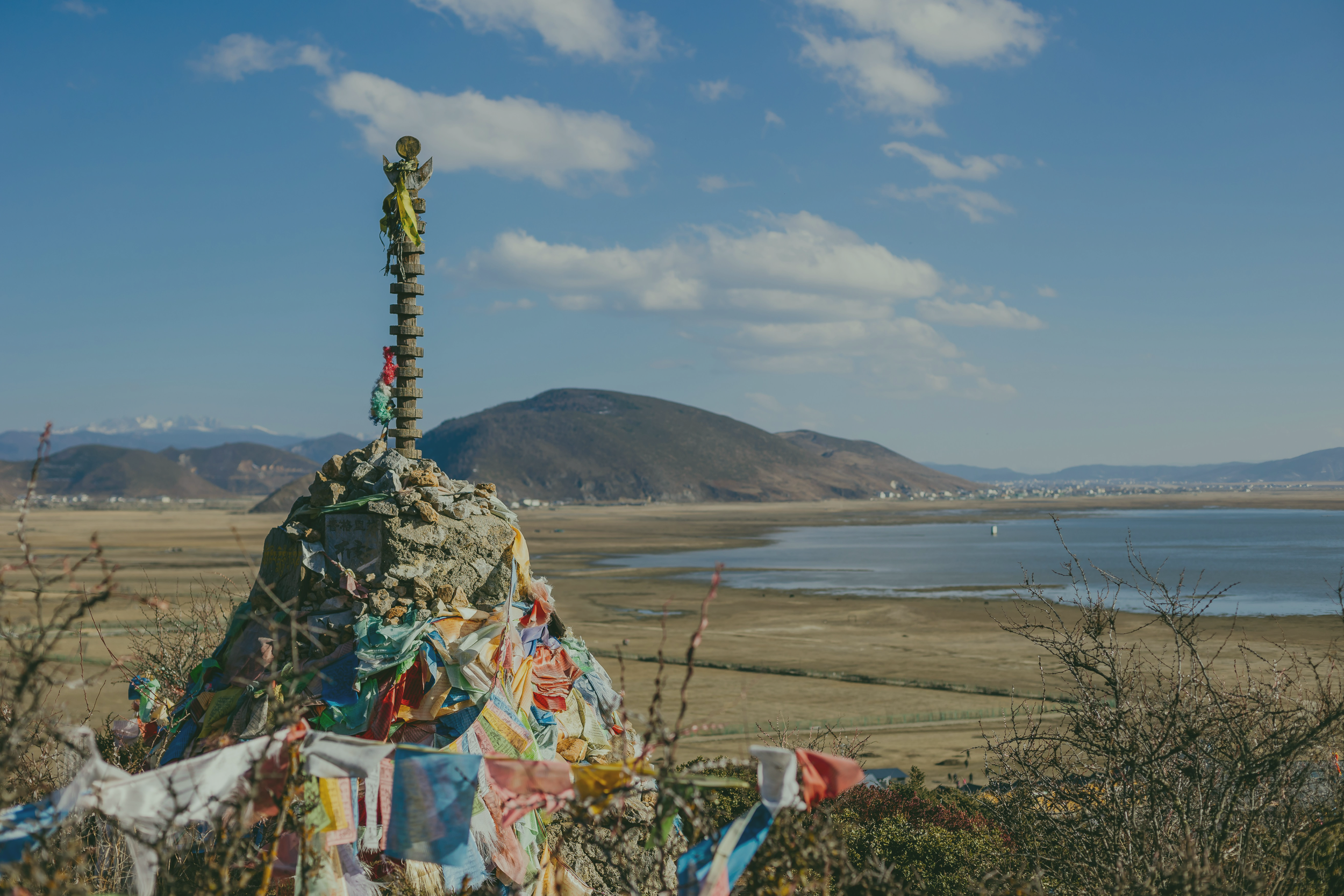 A rock cairn with colorful prayer flags and a figure at the top, overlooking a large river and mountains in the background.