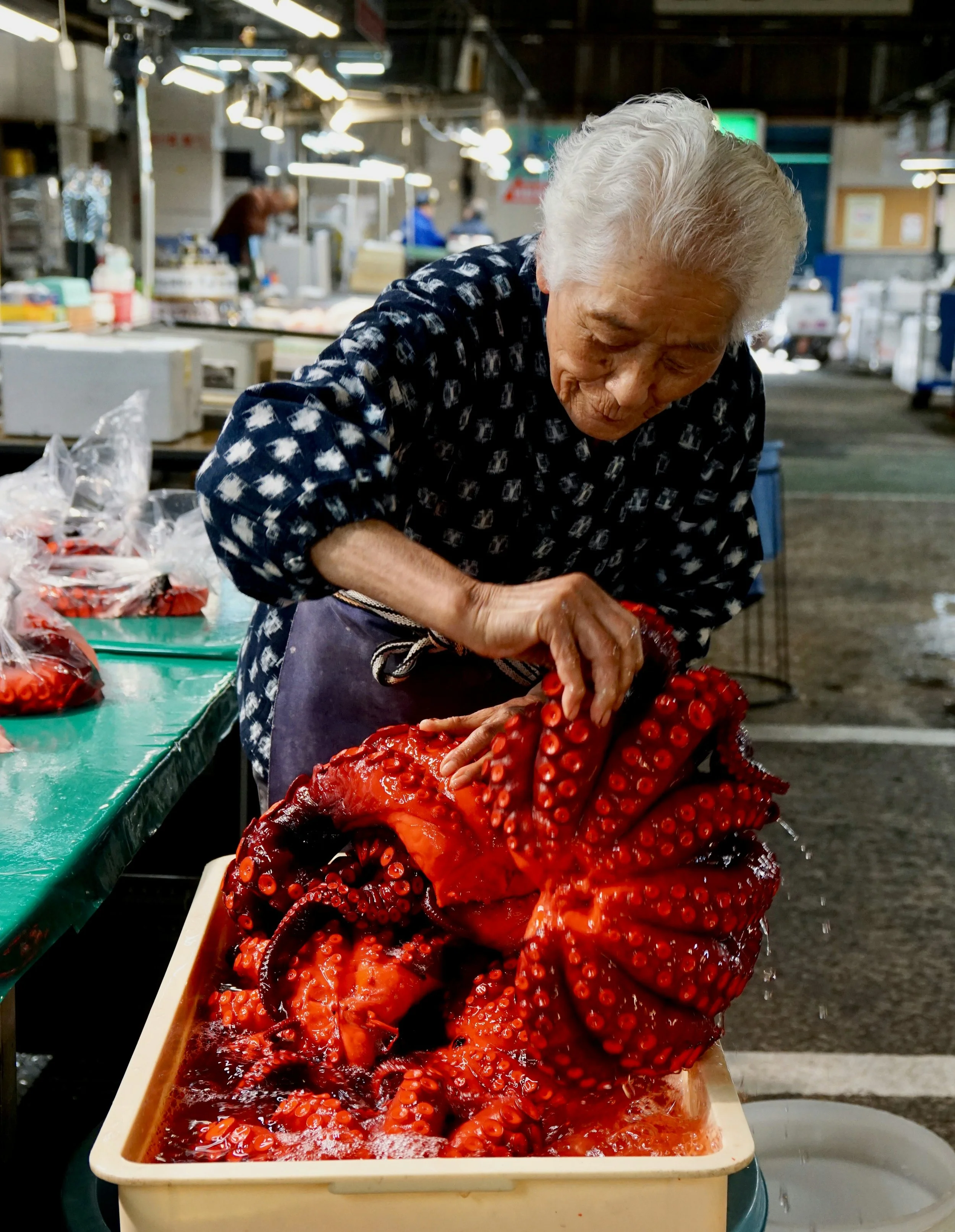 An elderly woman preparing octopus at a market stall in Japan, with the tentacles in a plastic container.