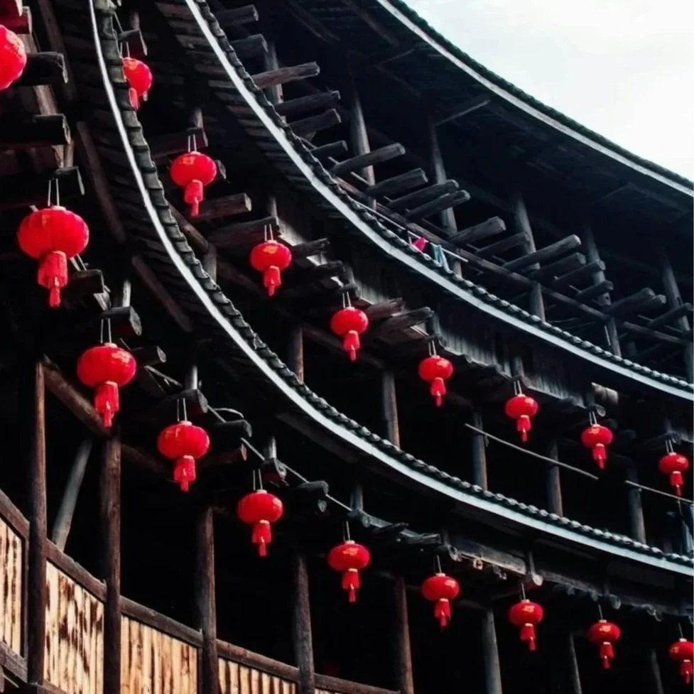 Traditional tulou wooden building decorated with red lanterns hanging along the eaves, under a partly cloudy sky in Fujian, China.