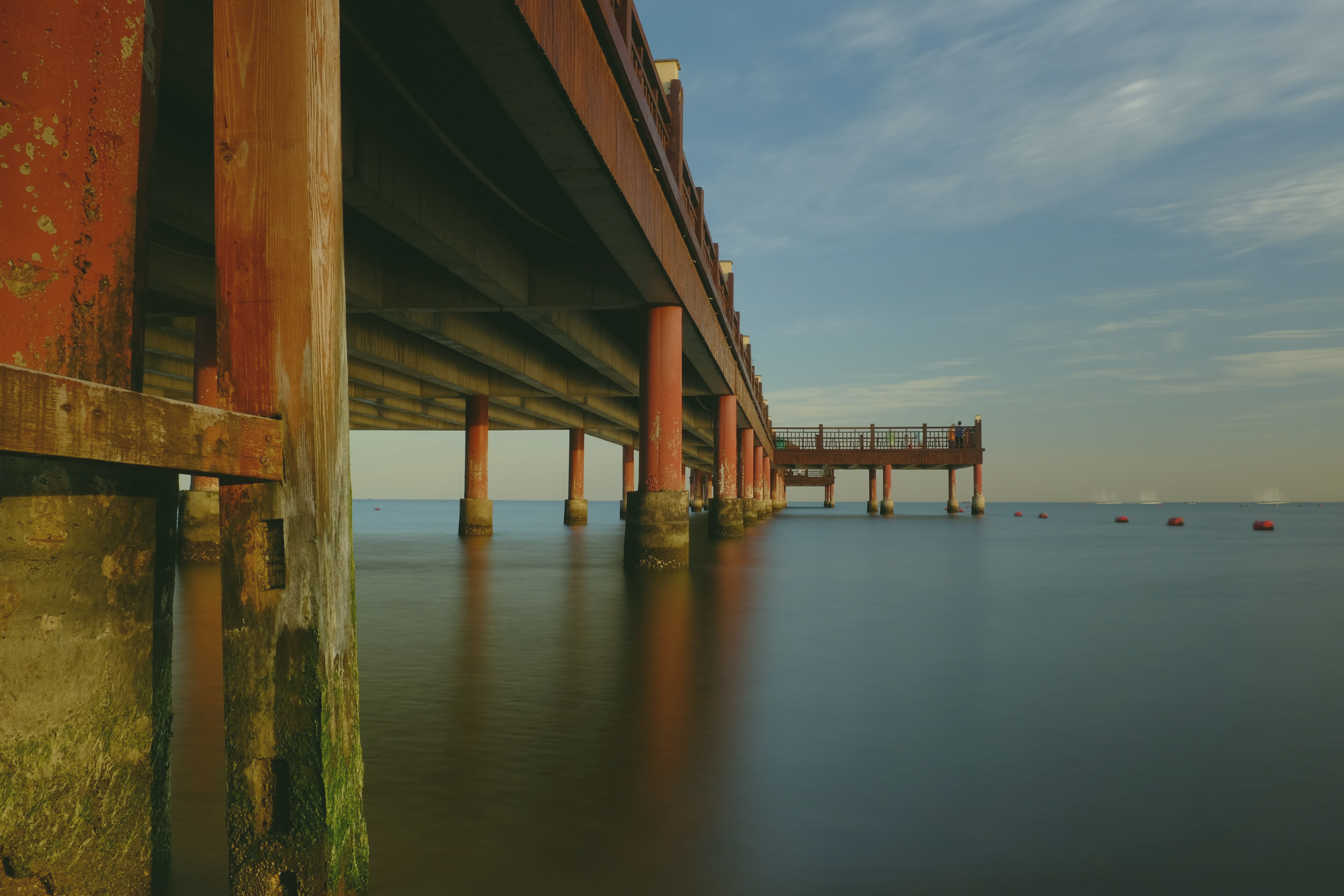 View of a pier extending over the ocean with red support columns, wooden railing, and a small observation area at the end, under a partly cloudy sky.