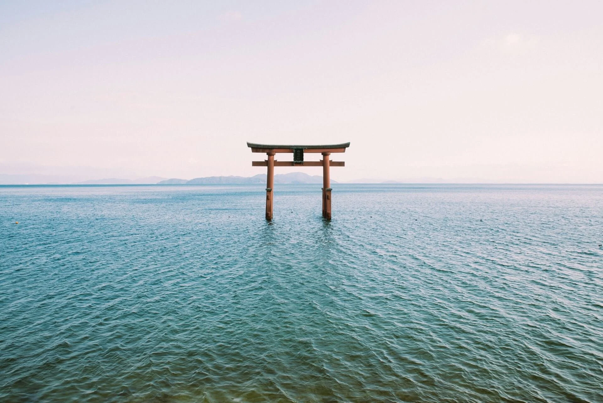 A traditional Japanese torii gate at Shirahige Shrine standing in the water of Lake Biwa with a distant horizon and mountains.