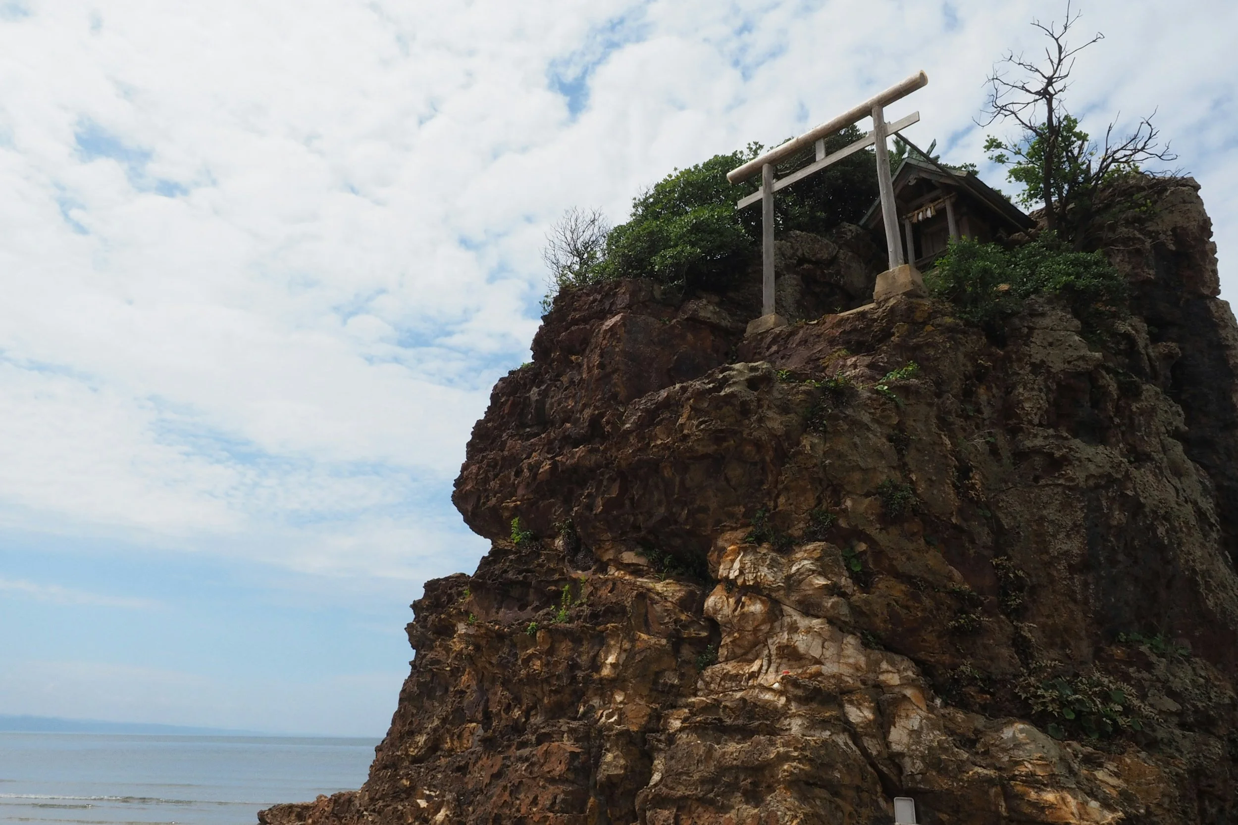 A rocky cliff with a small wooden structure and a torii gate on top, overlooking a calm body of water under a partly cloudy sky in Shimane, Japan.