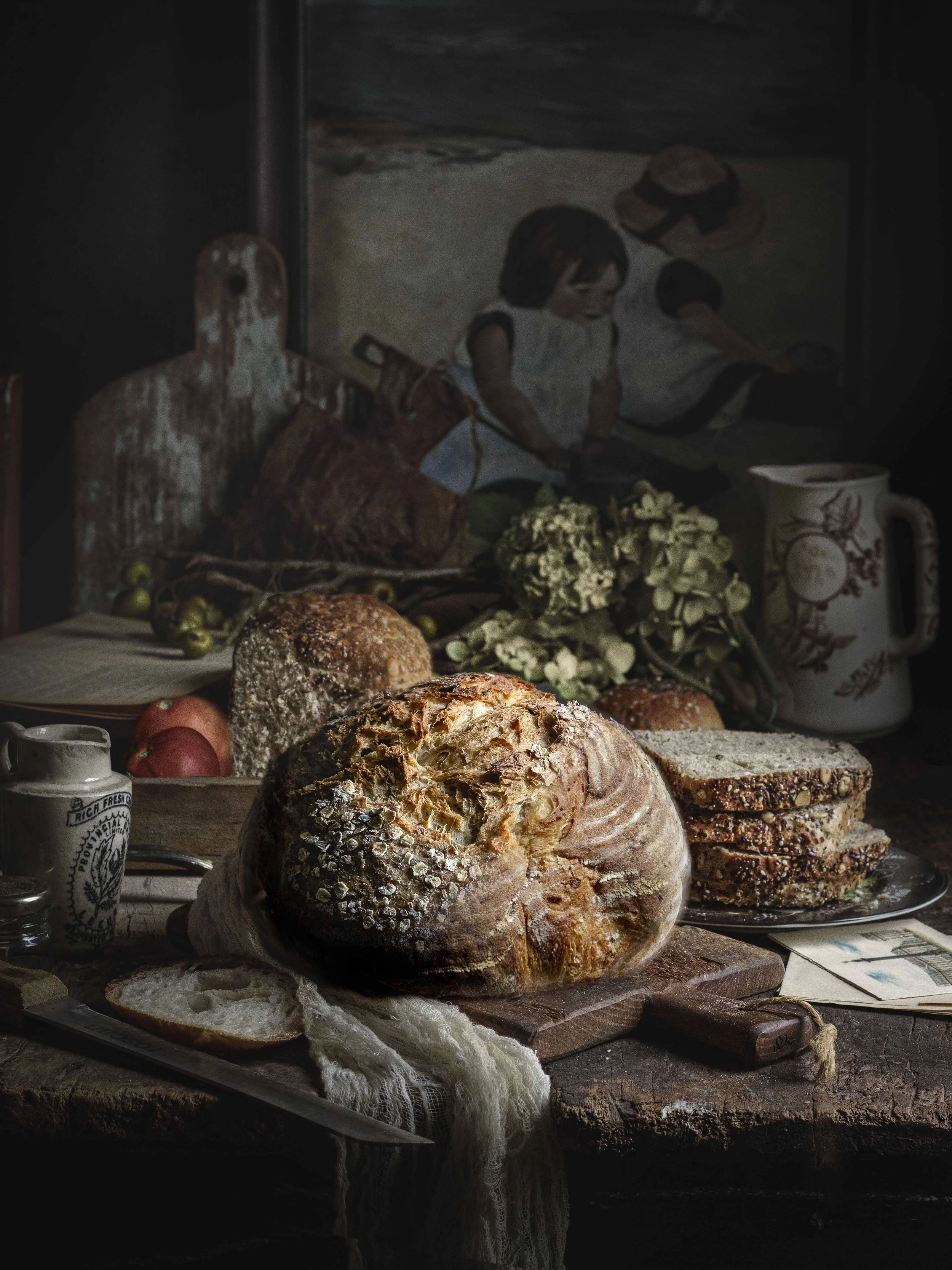 A rustic still life of bread on a wooden table, with a loaf in the foreground, sliced bread on a plate, and various other baked goods. The background features a painting of children and some vintage items, including a vase, flowers, and a book.