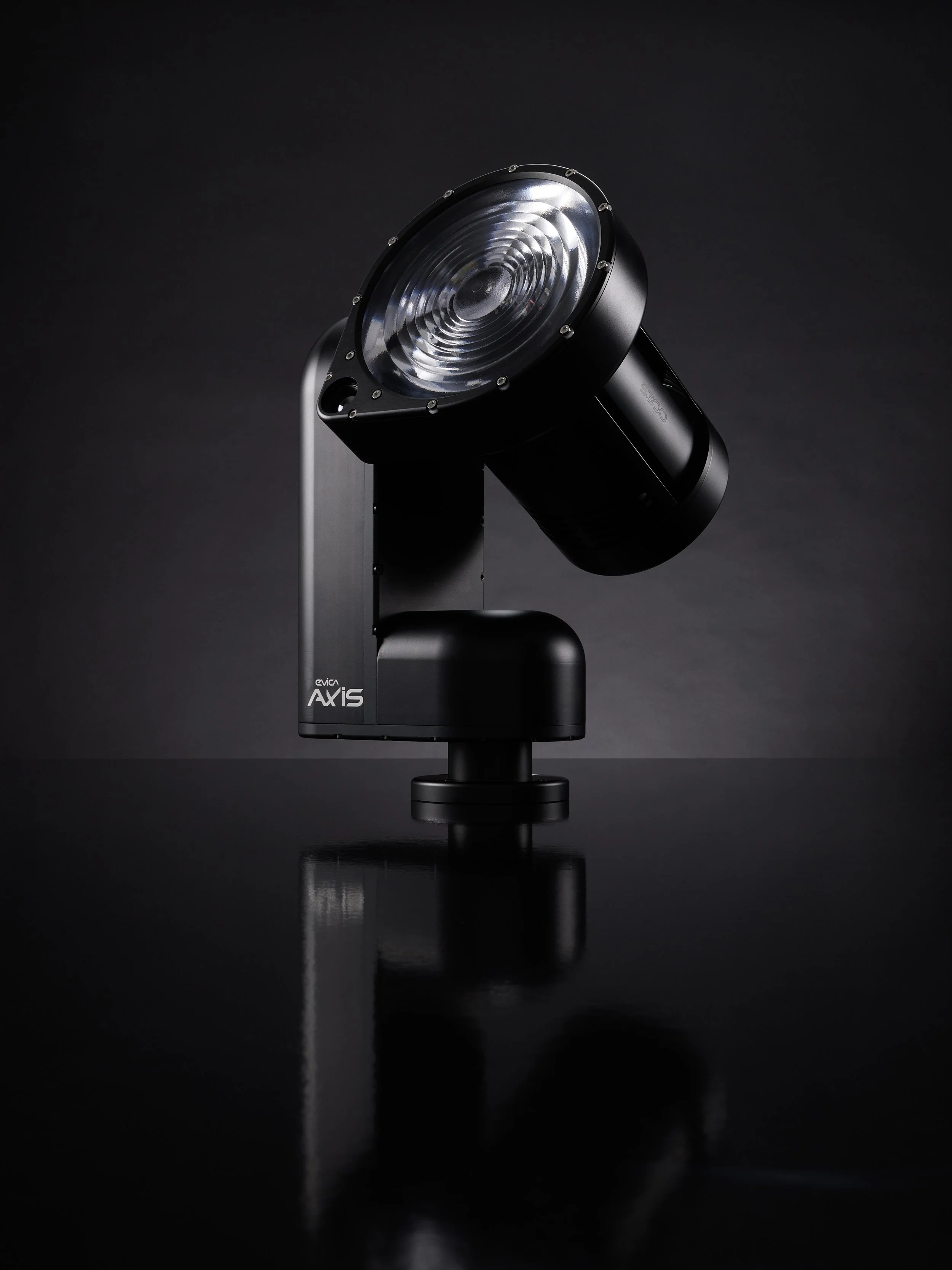 A black adjustable spotlight with a round reflective lens, mounted on a black base, reflected on a glossy surface against a dark background.
