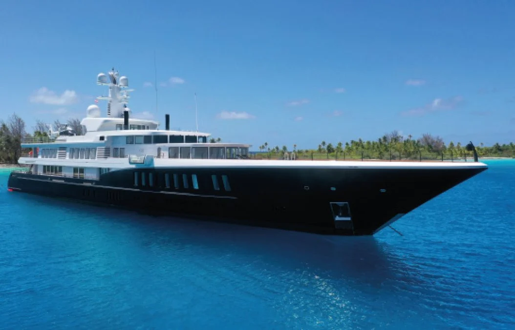 Large luxury yacht on calm blue water with a green island shoreline and partly cloudy sky in the background.