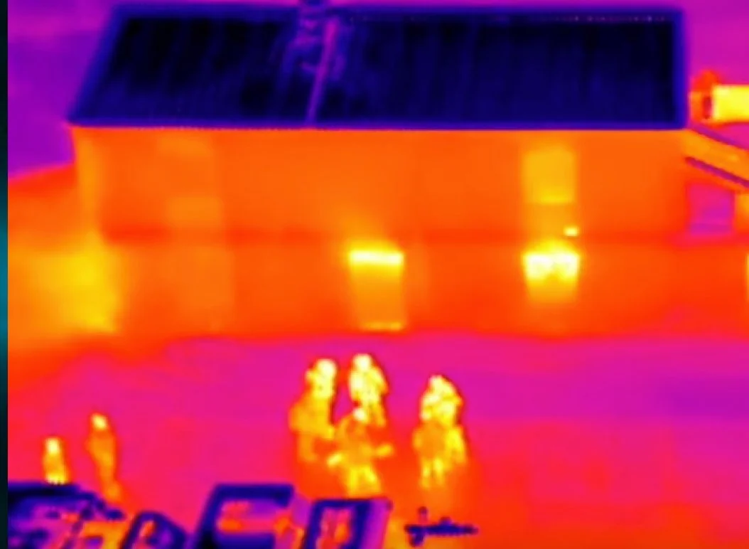 Infrared image of five individuals standing outside near a building, with the image showing heat signatures, where the people and the building emit different levels of heat.