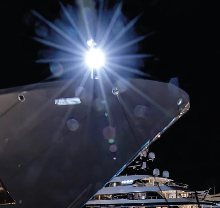 A bright light shines from the top of a large ship at night, with rays of light spreading outward, and part of the ship's upper deck and structures visible below.