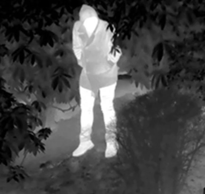 Person standing outdoors near bushes, wearing a jacket, shorts, and boots, with face obscured.